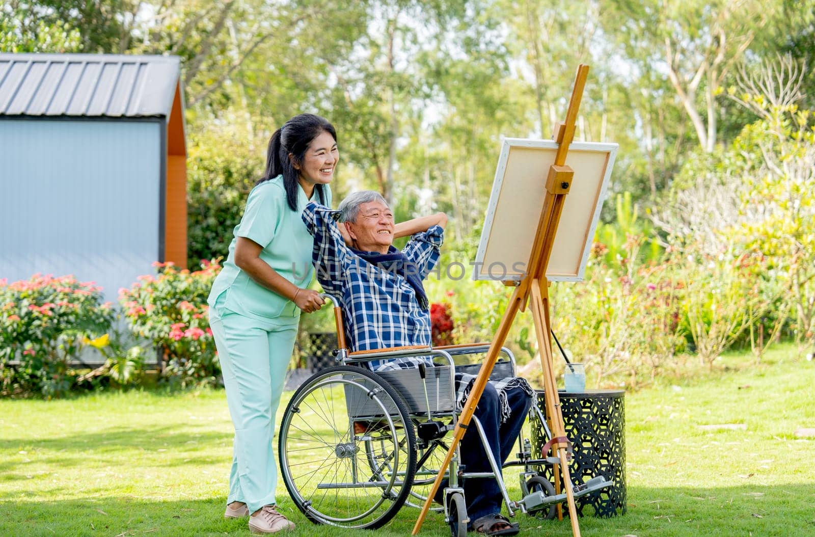 Asian senior man lean and relax action on wheelchair also look to painting after finish and nurse stay beside in the garden.