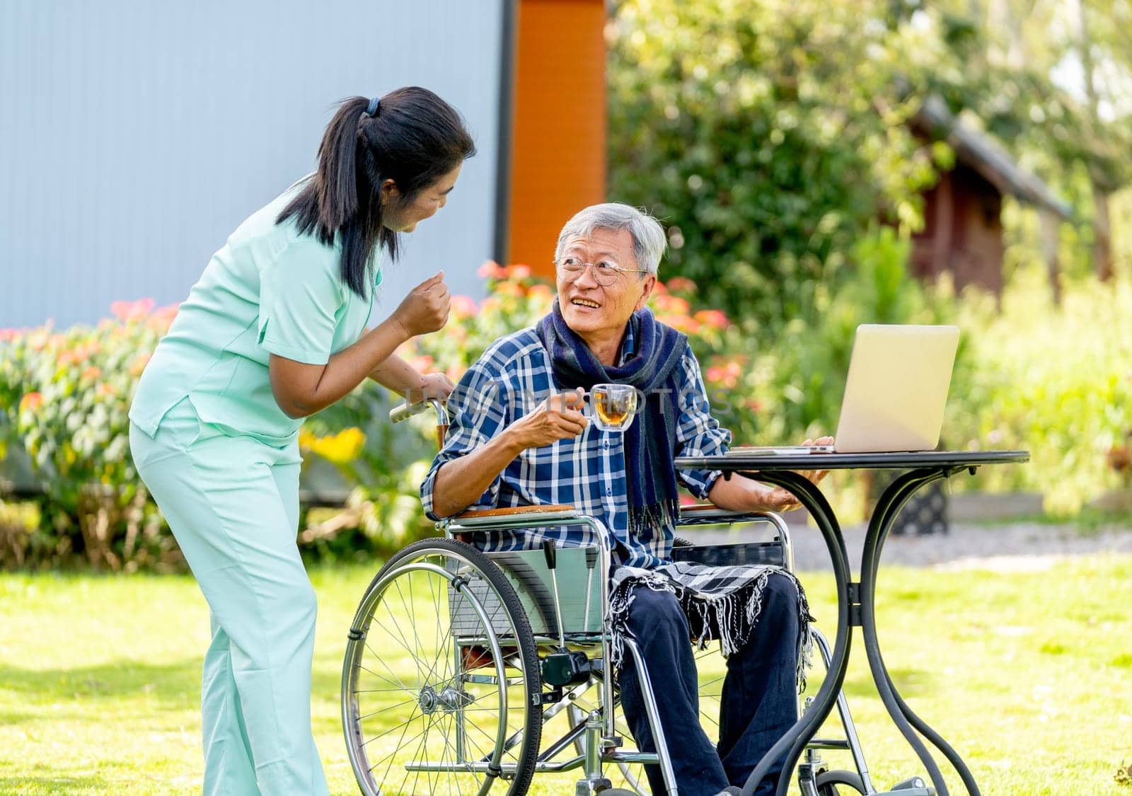 Side view of Asian nurse woman stand beside to take care senior man hold a glass of tea and sit on wheelchair also relax with laptop in the garden.