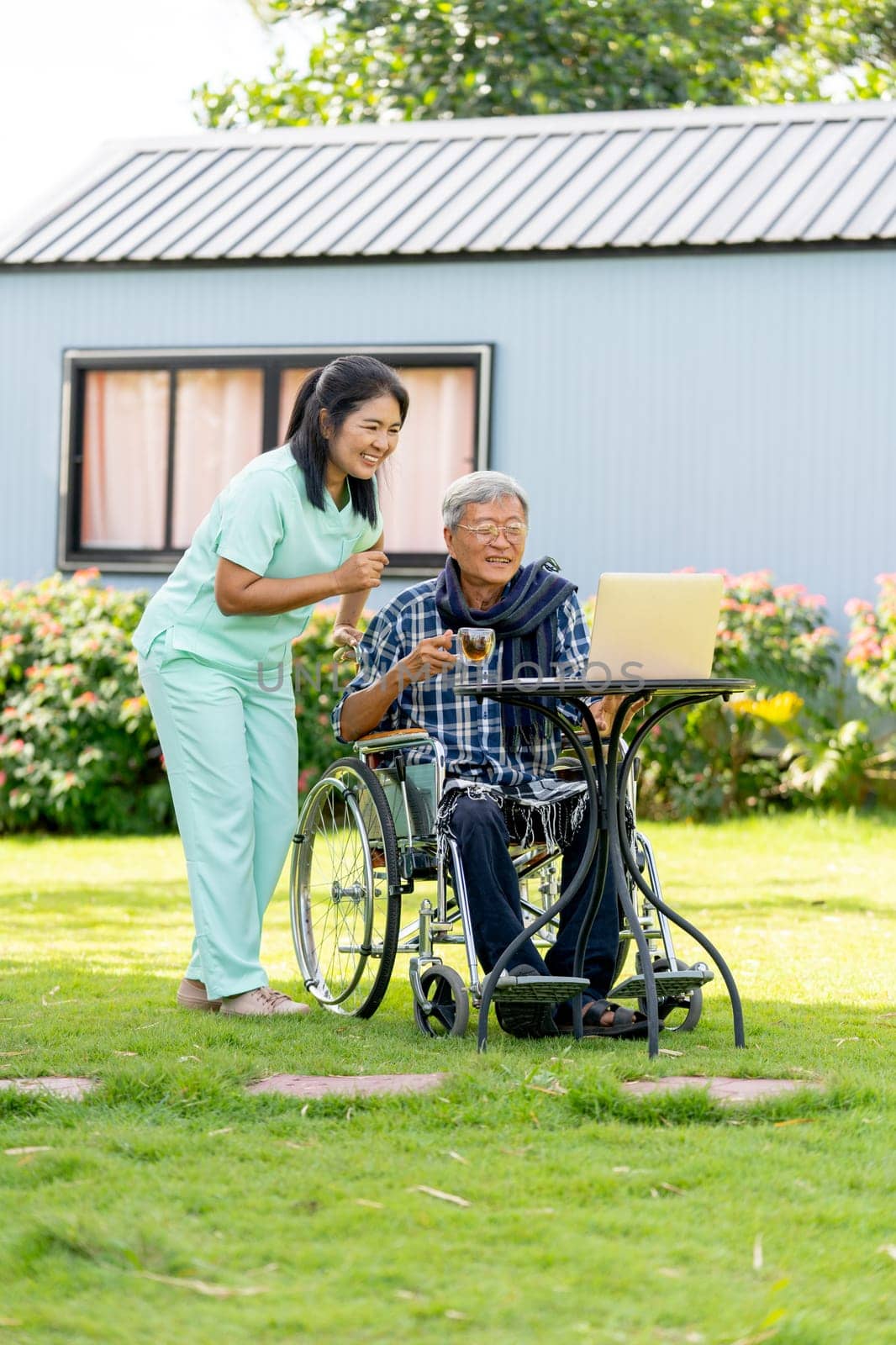 Vertical image of Asian nurse woman stand beside to take care senior man hold a glass of tea and sit on wheelchair also relax with watch the laptop in the garden.
