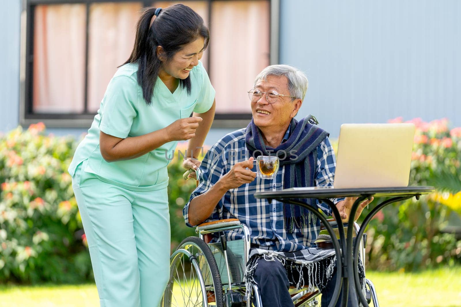 Asian nurse woman stand beside to take care senior man hold a glass of tea and sit on wheelchair also stay to relax in the garden.