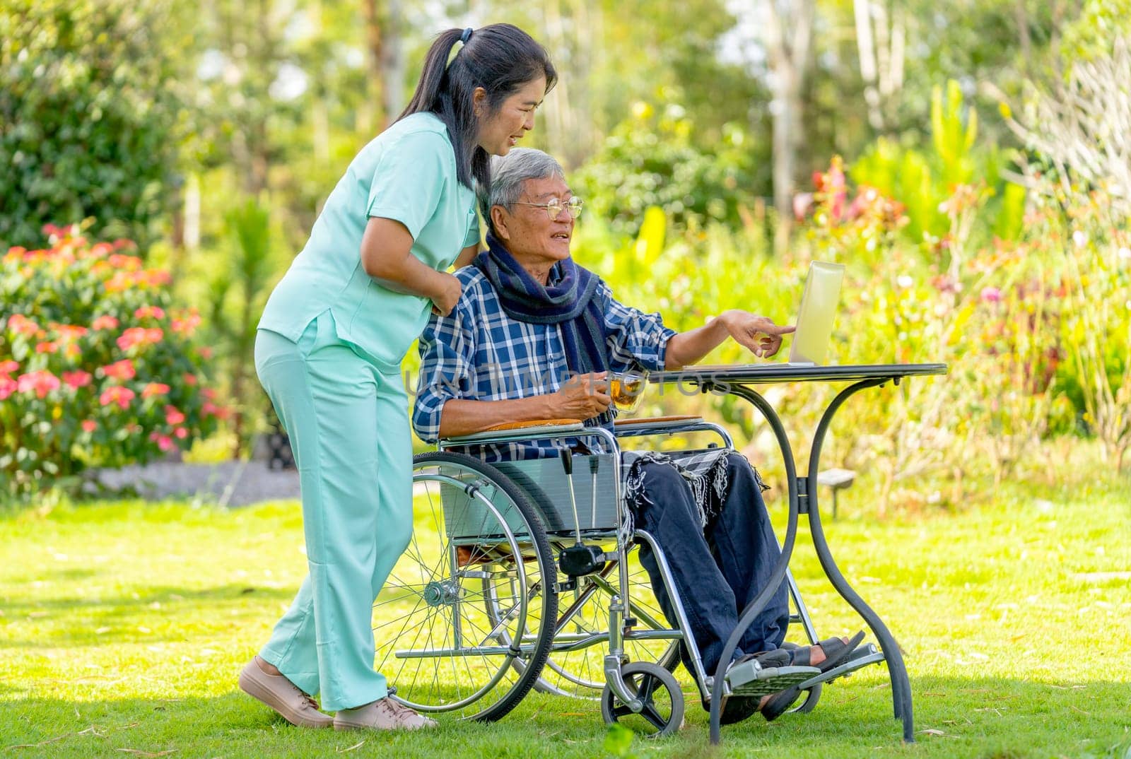 Side view of senior man on wheelchair point to monitor of laptop and talk with Asian nurse who take care him to get good health outdoor.