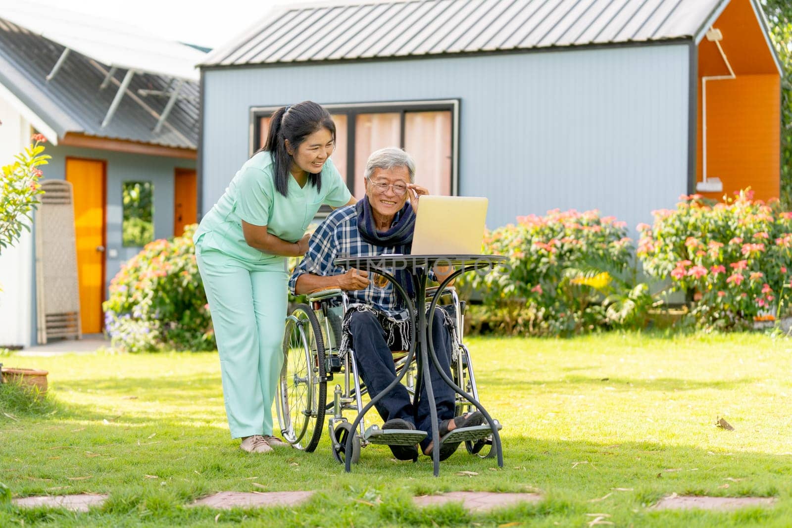 Wide shot of Asian nurse woman stand beside to take care senior man touch glassess and sit on wheelchair also stay to relax with laptop in the garden.