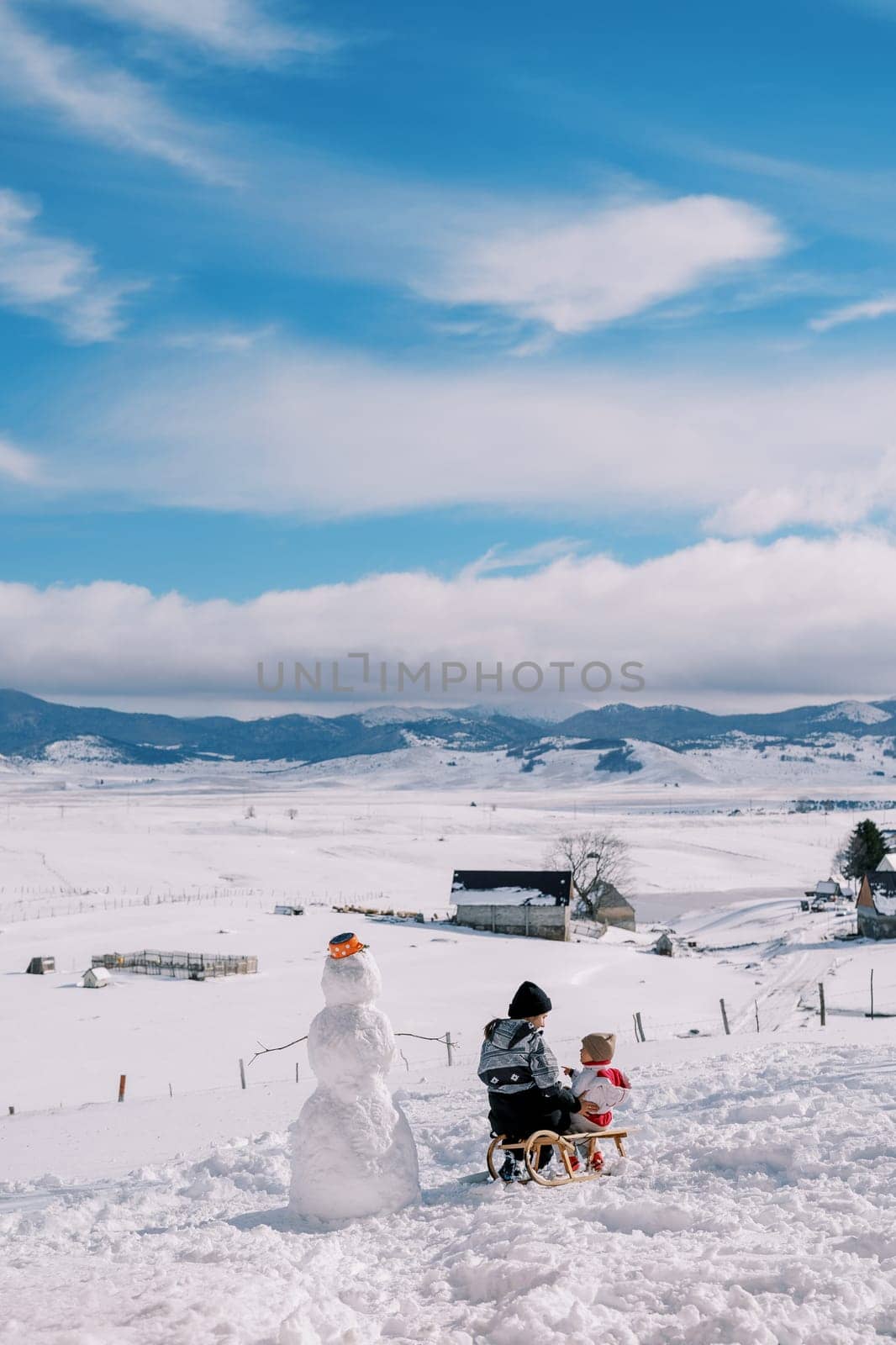 Mom and little girl are sitting on a sled near a snowman and look at each other. Back view. High quality photo