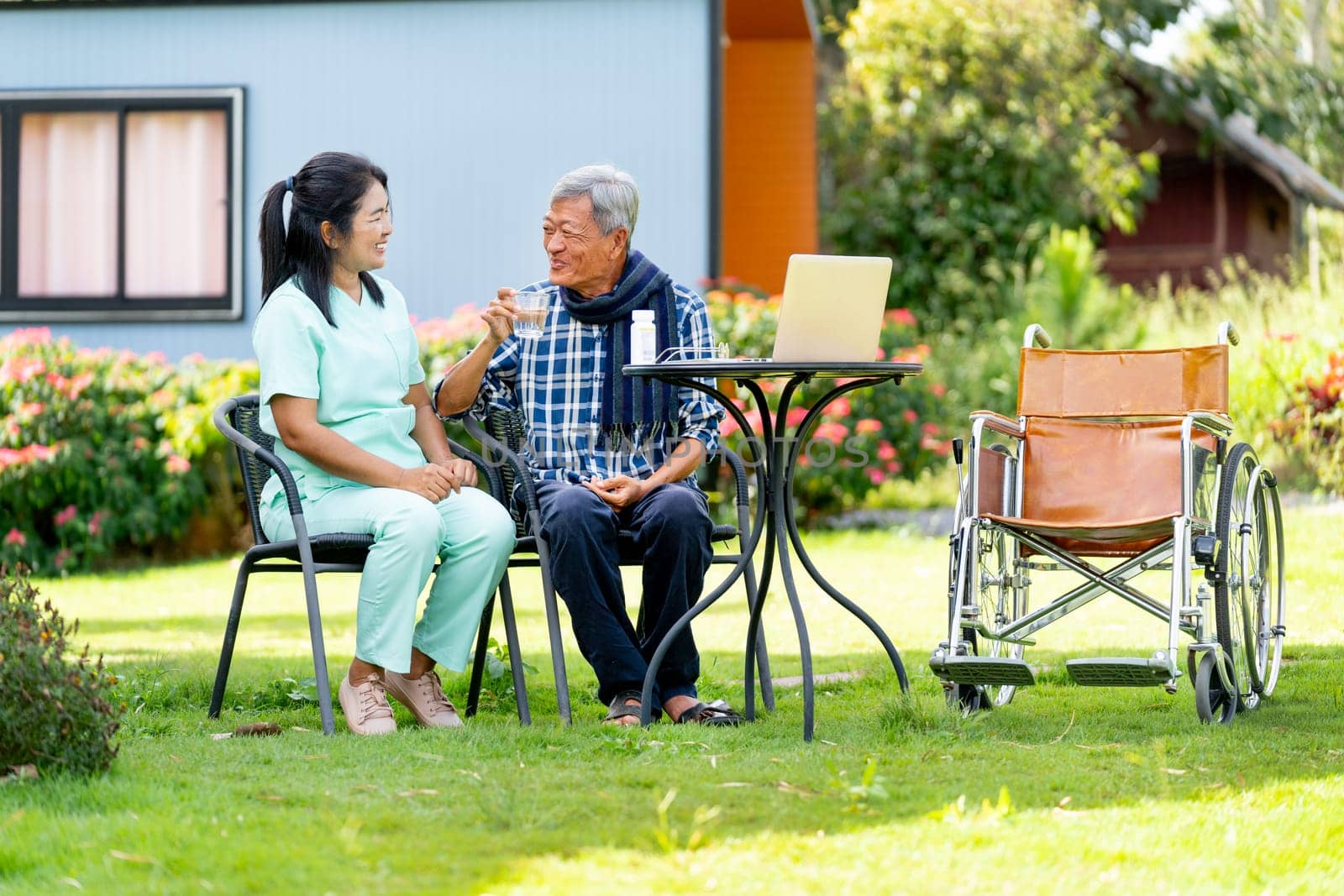 Asian senior man drink water from glass after take the medicine from nurse and they sit outdoor in the garden with wheelchair is set beside.