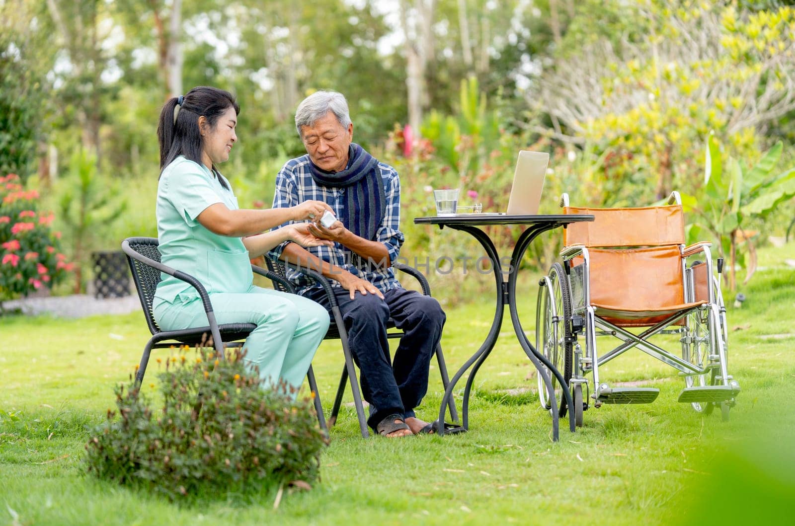 Asian nurse woman give the medicine pill from bottle to senior man and they sit on chair in the garden with wheelchair is set beside.
