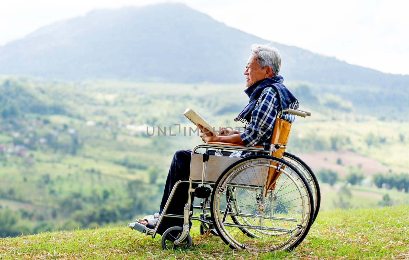 Asian senior man sit on wheelchair and read book also stay near cliff with meadow and mountain in the background with warm light and concept of nature health care.
