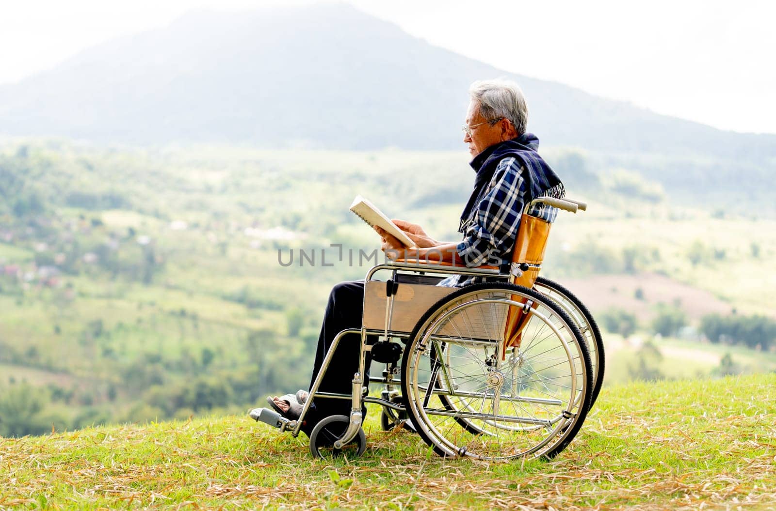 Asian senior man with eyeglasses read the book and sit on wheelchair also stay near the cliff with grassfield and mountain as background.
