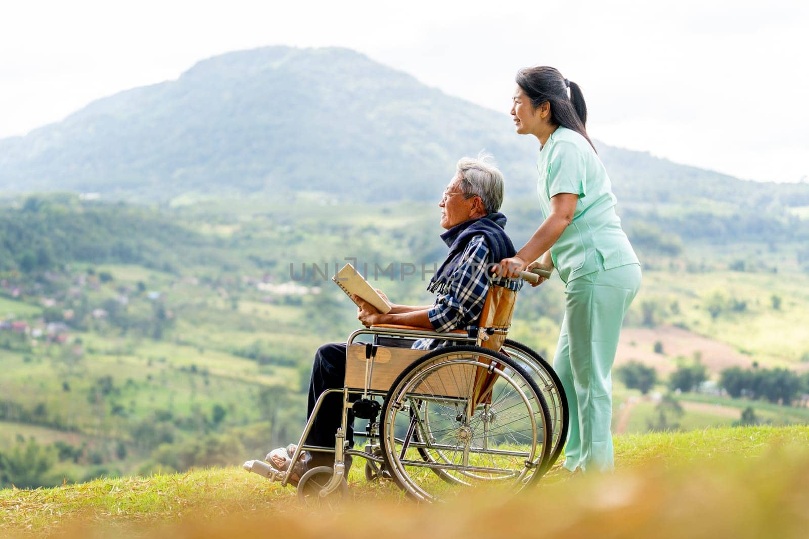 Asian nurse stand behide and hold wheelchair handle with senior man sit on wheelchair and both look forward and stay near cliff with village and mountain in the background.