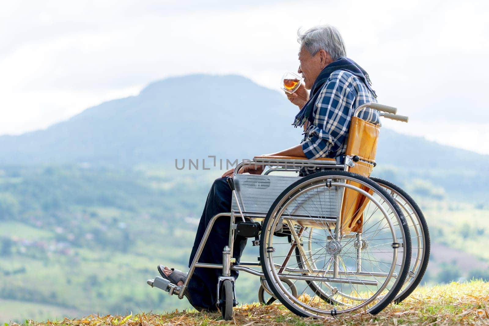 Asian senior man drink tea from a cup and sit on wheelchair with near the cliff and mountain and grassfield as background.