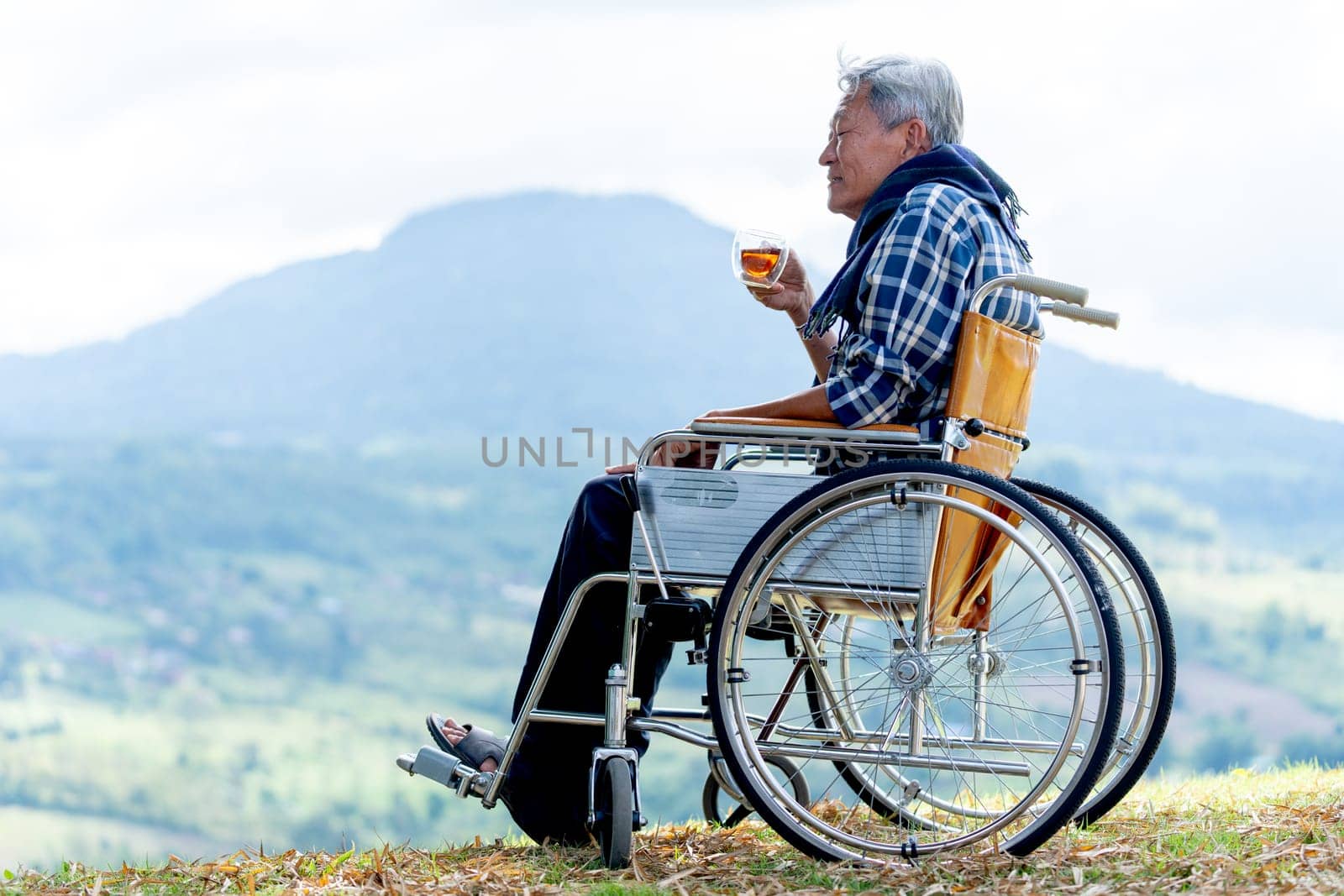 Asian senior man hold a cup of tea and sit on wheelchair with lood forward and stay near the cliff and mountain and grassfield as background.