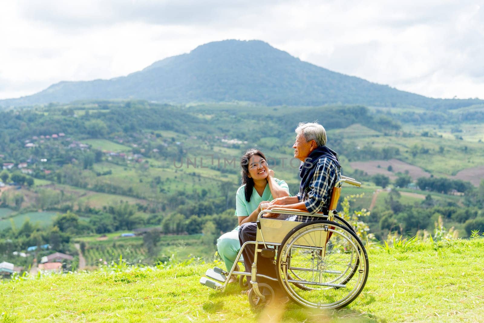 Asian nurse sit near senior man on wheelchair to take care for good health outdoor and both stay near the cliff with warm light.