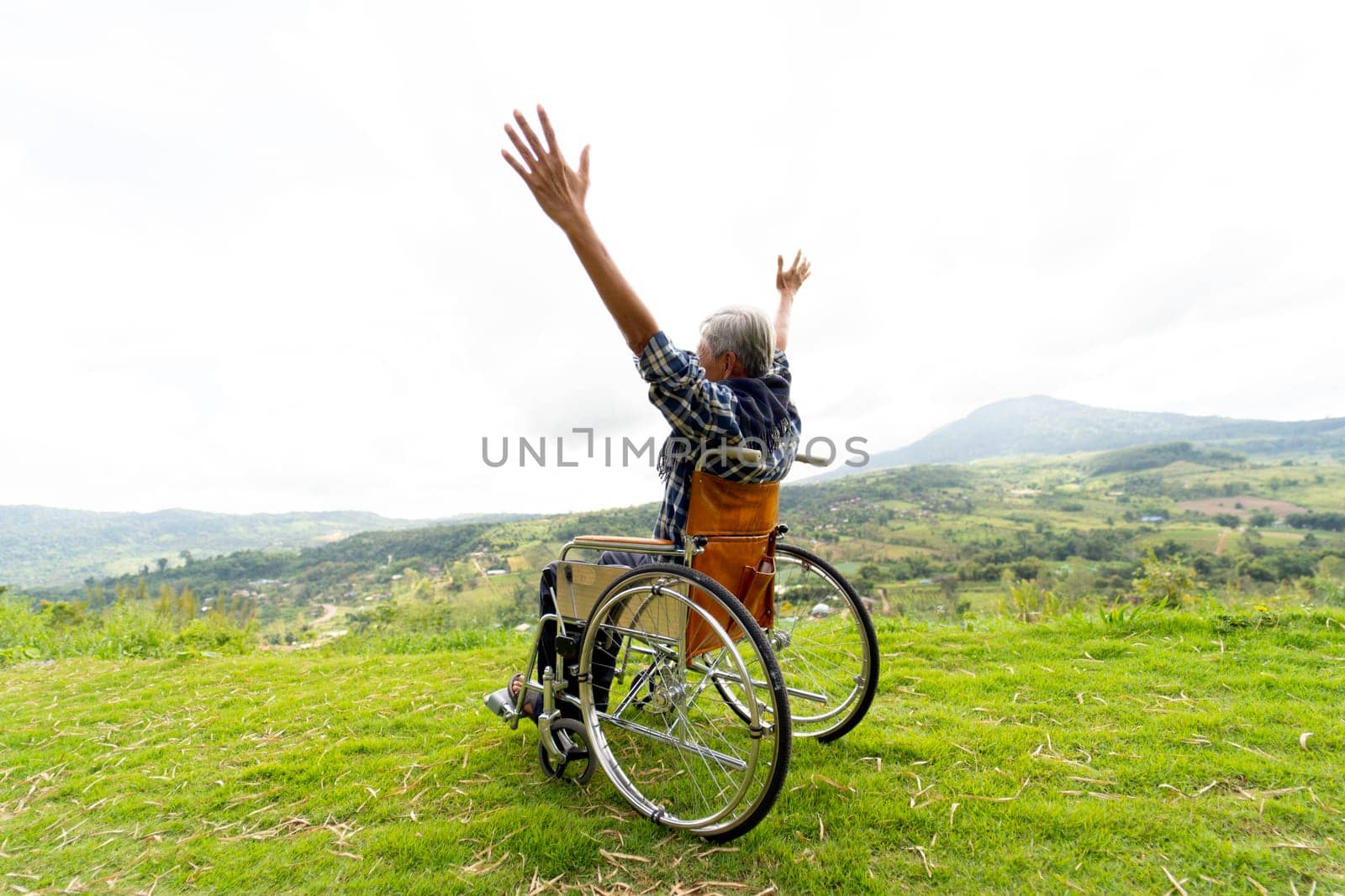 Back of senior man sit on wheelchair and action of raise his hands up like stretching also stay near cliff with mountain and grass field in the background.