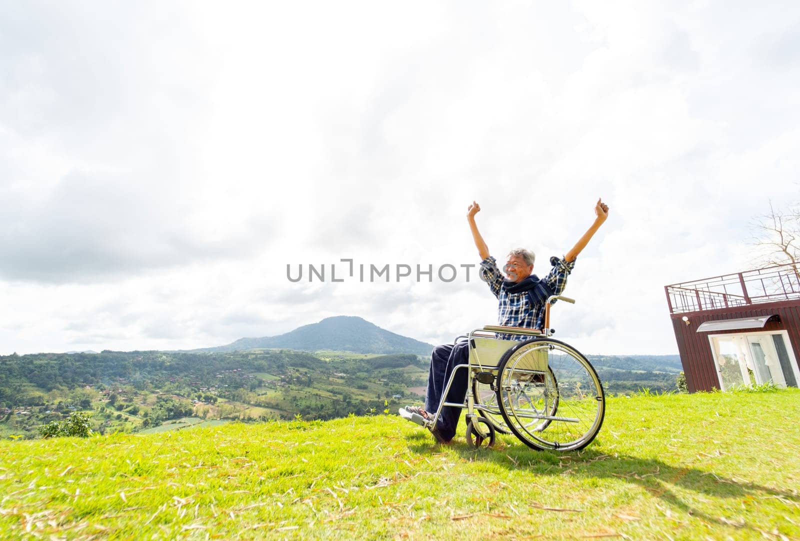 Wide shot of Asian senior man sit on wheelchair and action of raise his hands up like stretching also stay near cliff with mountain and grass field in the background.