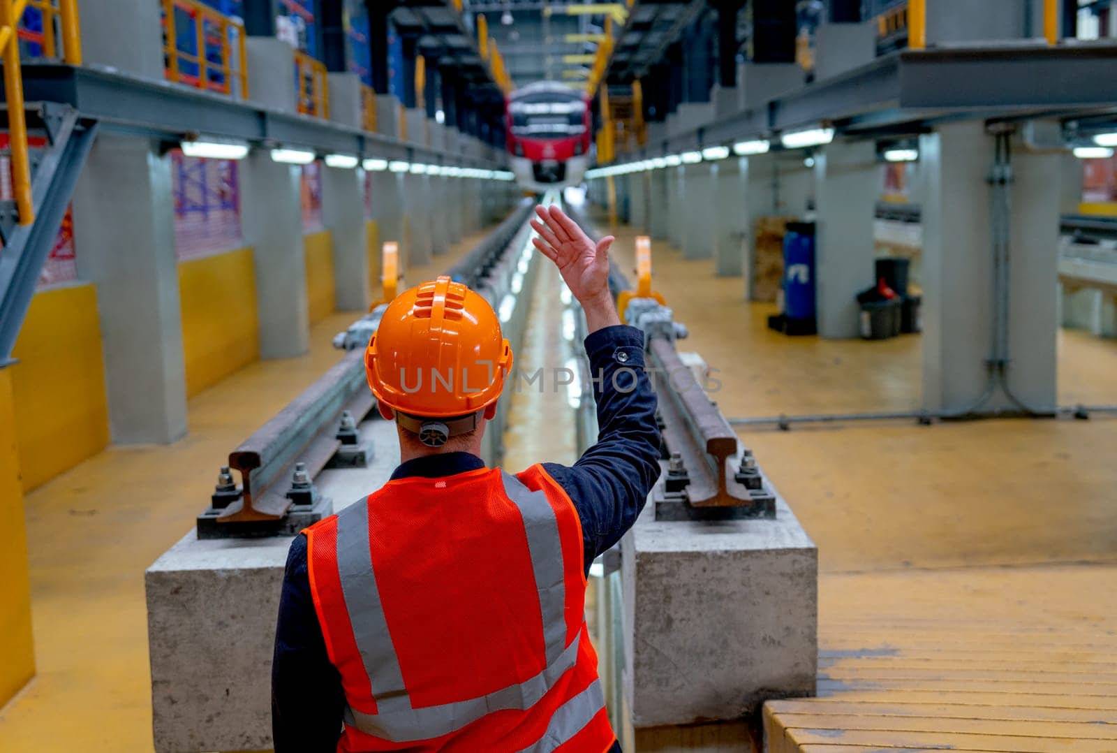 Back of technician worker with safety uniform show body signal to electrical or metro train and stand in front of railroad tracks in factory workplace.