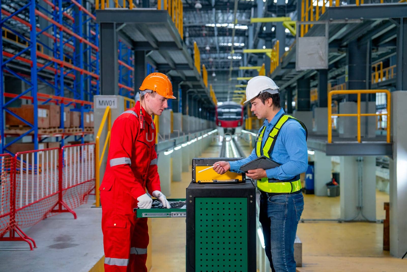 Technician and engineer worker discuss about tools and equipment with cabinet in front of railroad tracks of electrical or sky train in factory workplace.