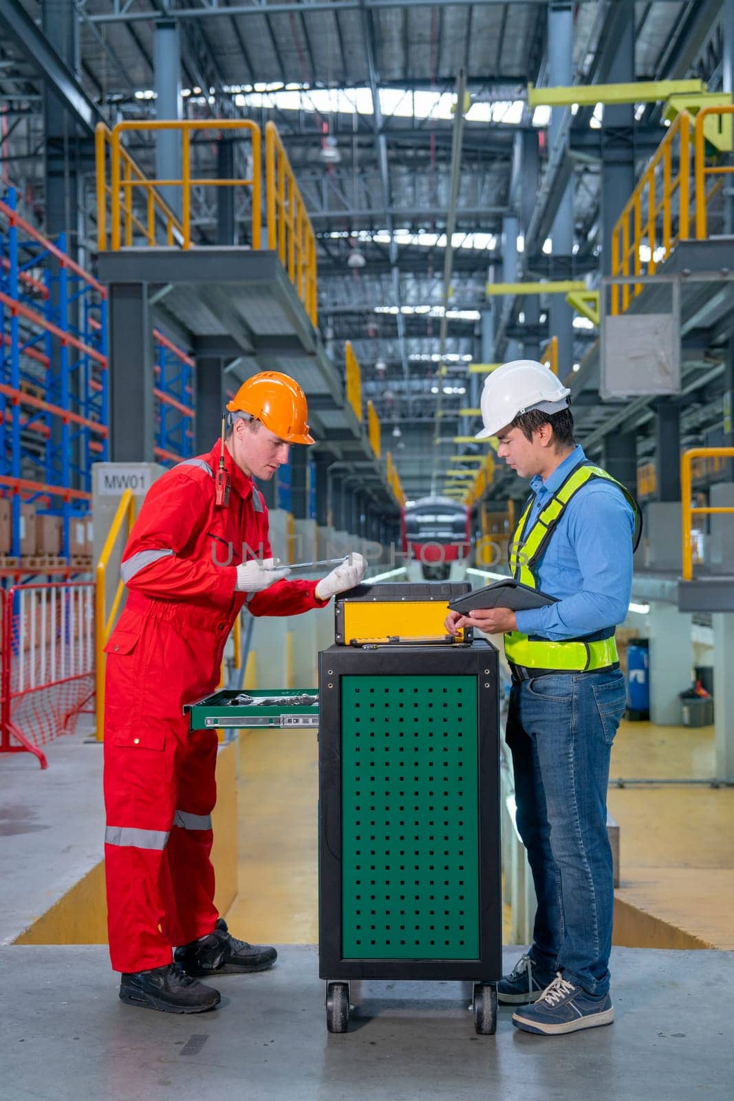 Vertical image of technician and engineer workers discuss about tools and equipment with cabinet in front of railroad tracks of electrical or sky train in factory workplace.