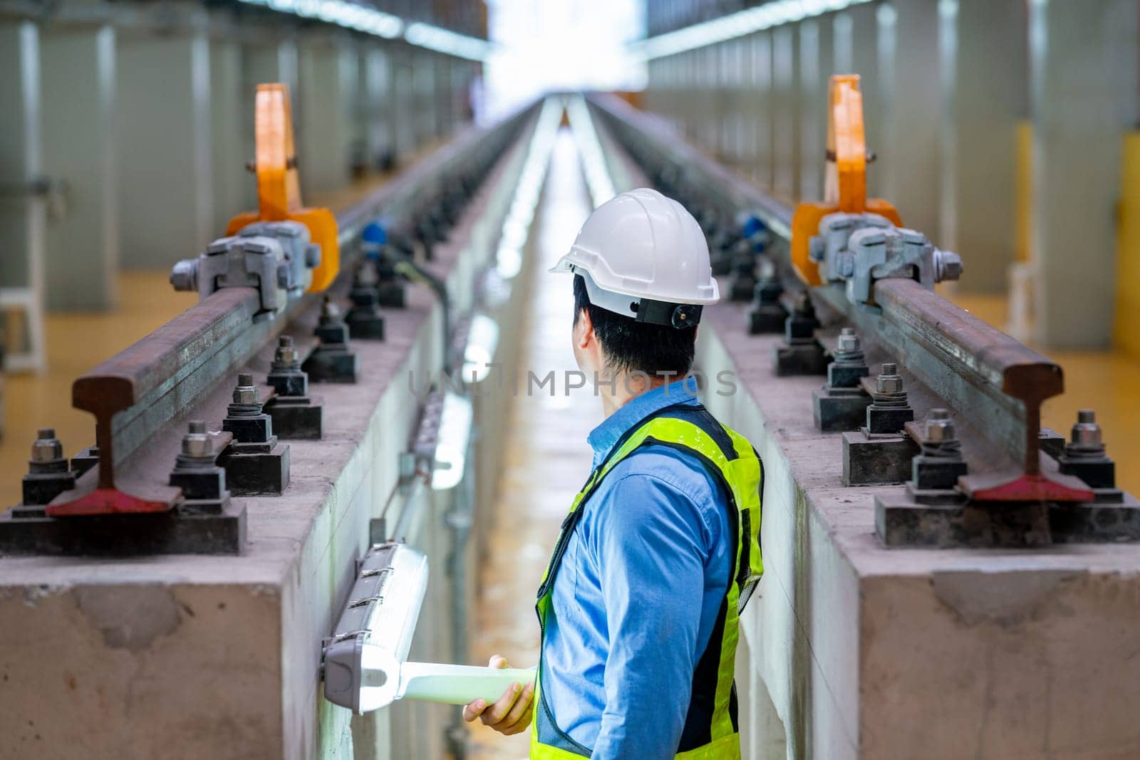 Back of professional engineer stand between railroad tracks of electrical or sky train and look to end of the rail in the factory workplace.