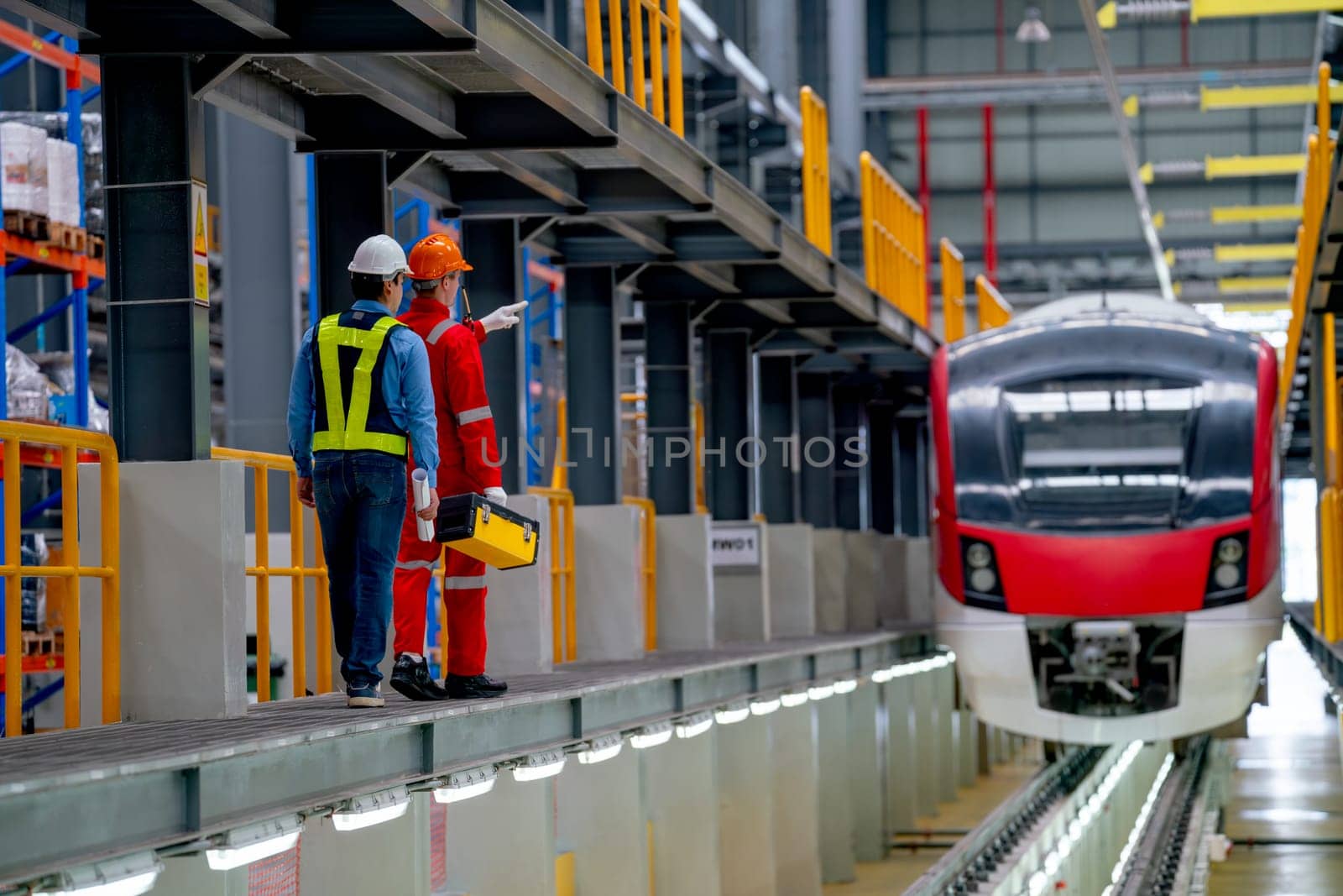 Back of engineer and technician workers bring tool box and walk beside railroad tracks of electrical or sky train in factory workplace and go to check and maintenance the train.