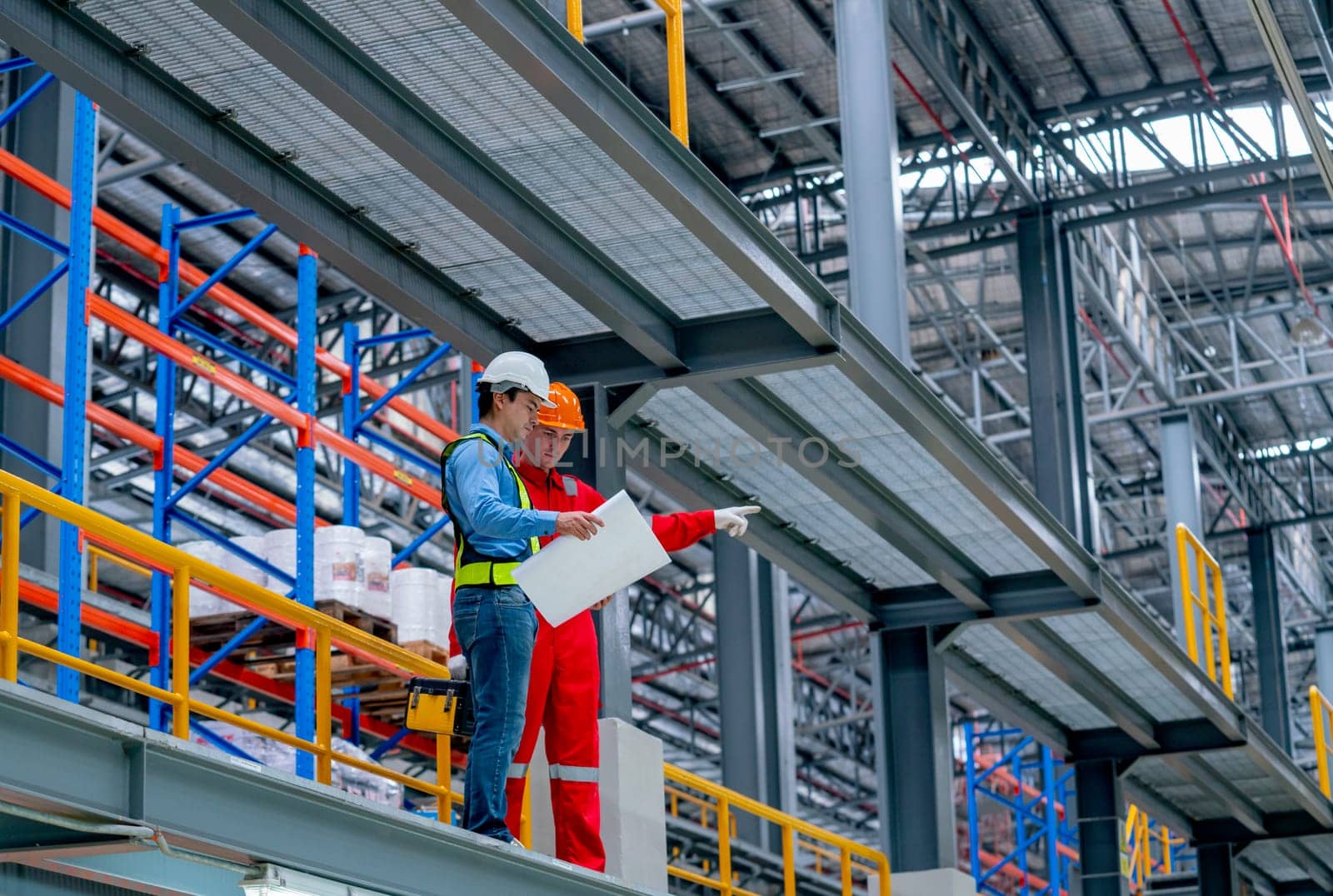 Professional engineer hold drawing plan and technician worker stay and discuss beside railroad tracks in the factory. The technician also point to left side to explain to his coworker.