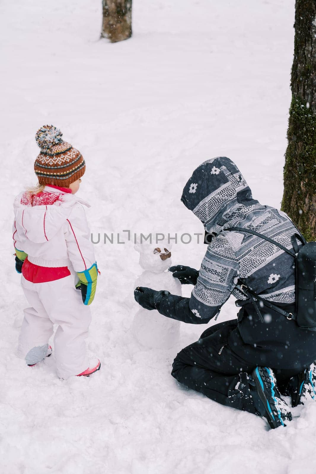 Little girl looks at a snowman made by her mother in the forest. High quality photo