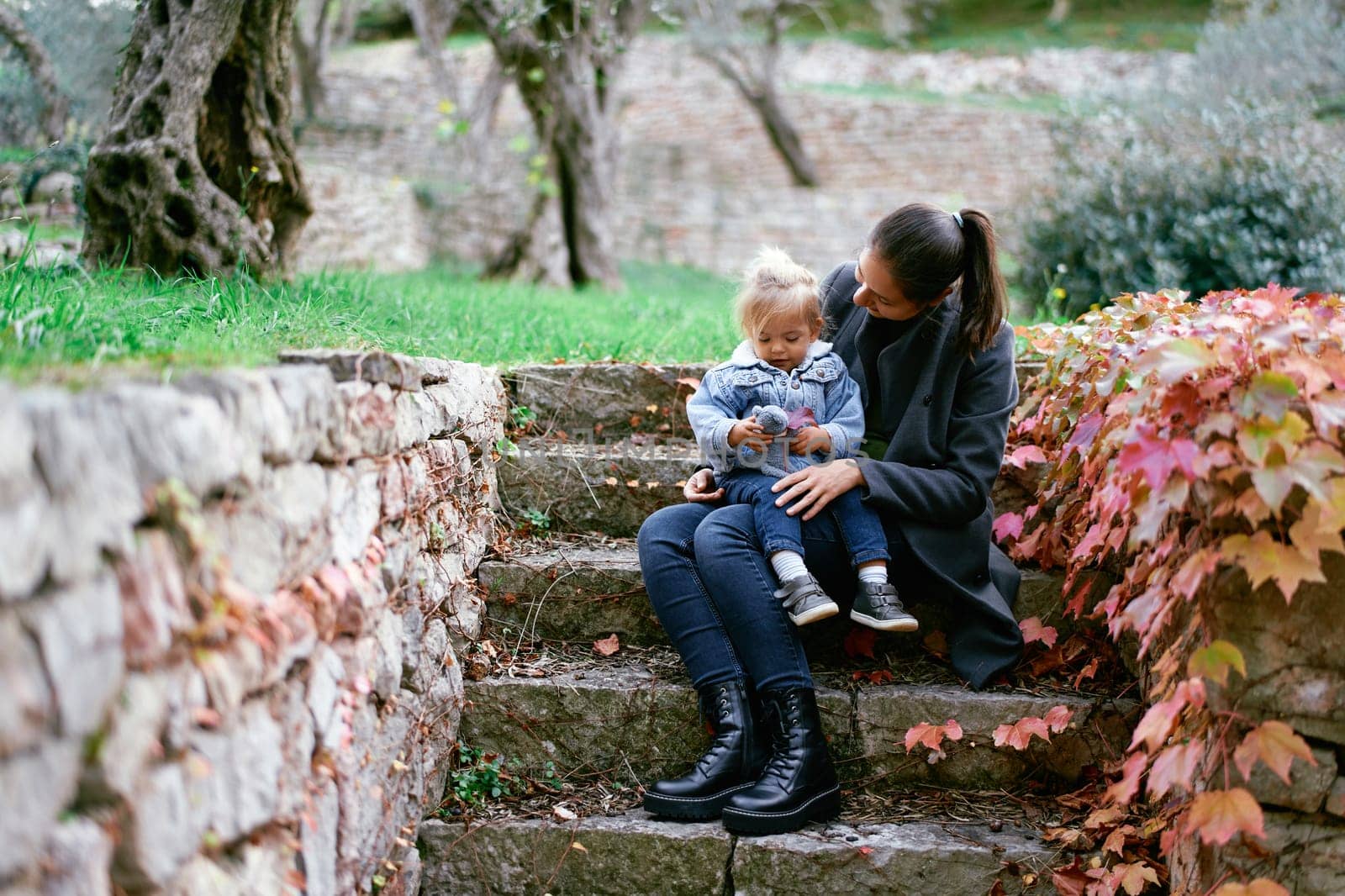 Mom with a little girl on her knees sits on stone steps in the park and looks at her. High quality photo