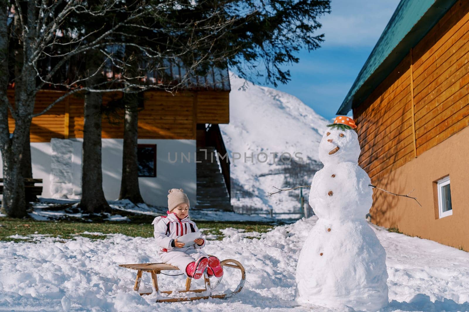 Little girl sits on a sled near a snowman next to a wooden house. High quality photo