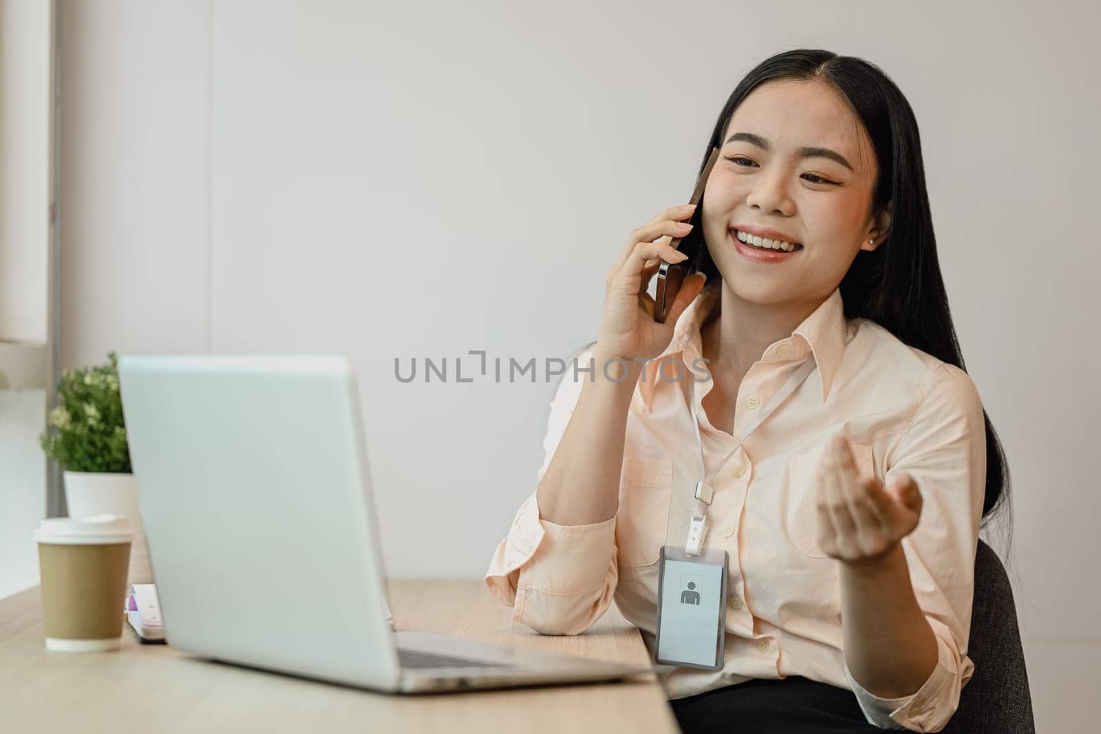Attractive Asian female employee talking on mobile phone and using laptop at desk.