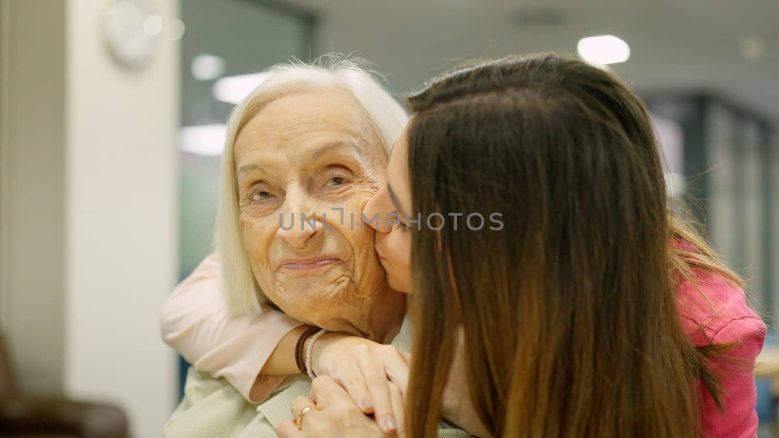 Smiling nurse that is a granddaughter kissing and embracing a senior woman in geriatrics