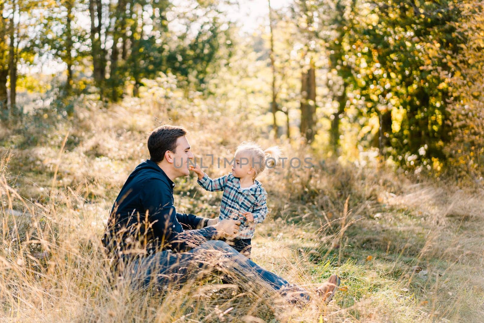 Little girl touches the face of her dad sitting on the lawn. Side view by Nadtochiy