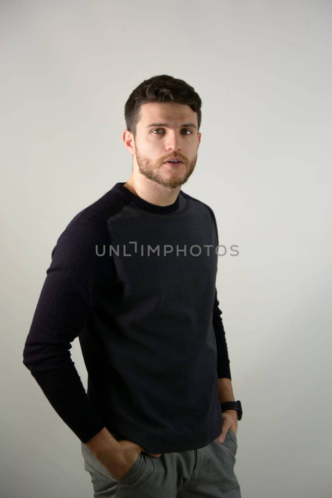 Attractive young man with blue eyes, wearing black elegant sweater, in studio shot on neutral background with hands in pockets