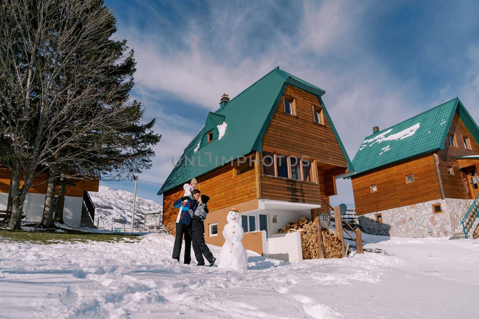 Mom kisses dad with little girl on his shoulders standing next to snowman near wooden chalet. High quality photo