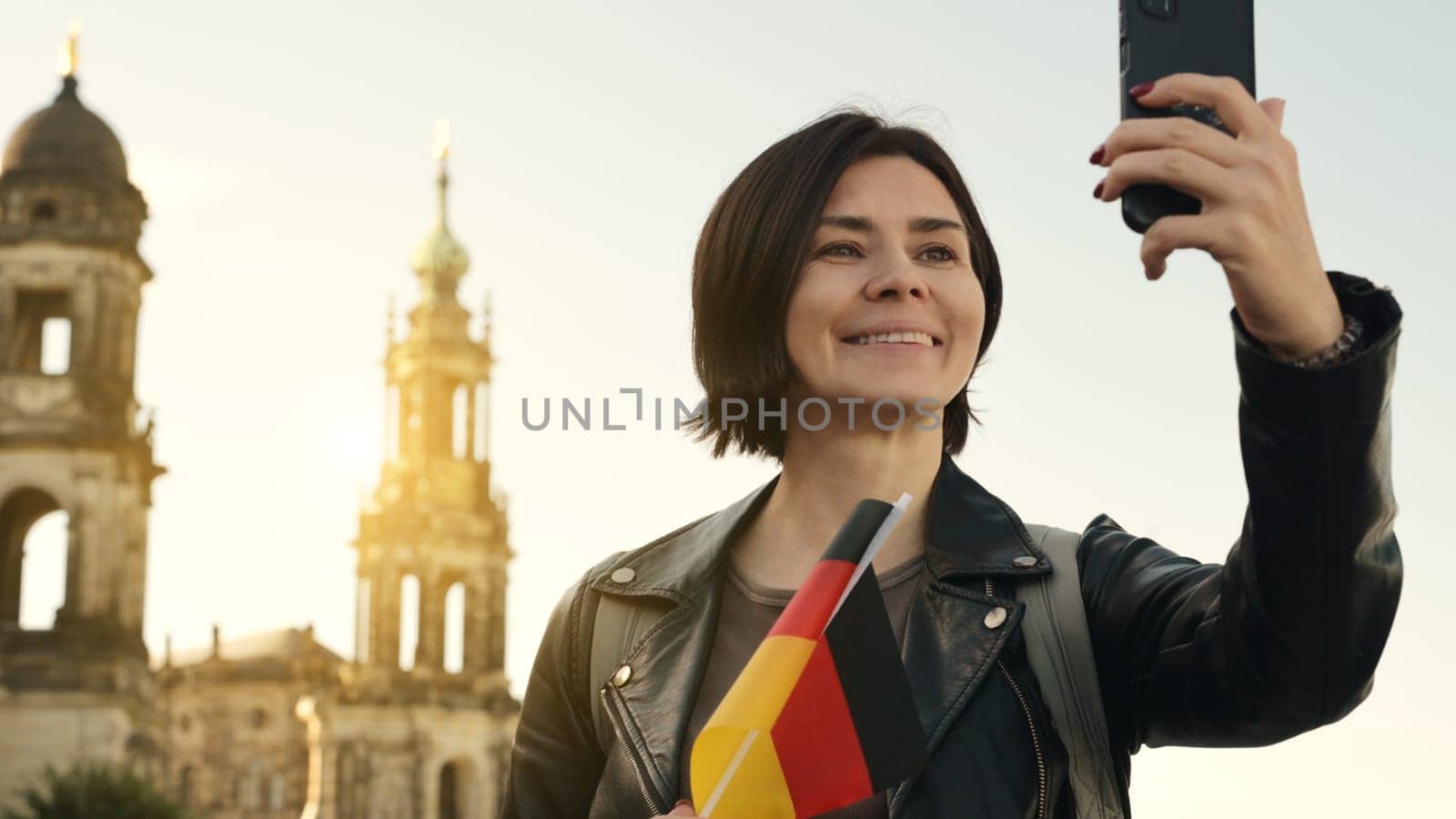Young Woman Takes Selfie With German Flag In Hand, Against Blurred City Backdrop In Autumn