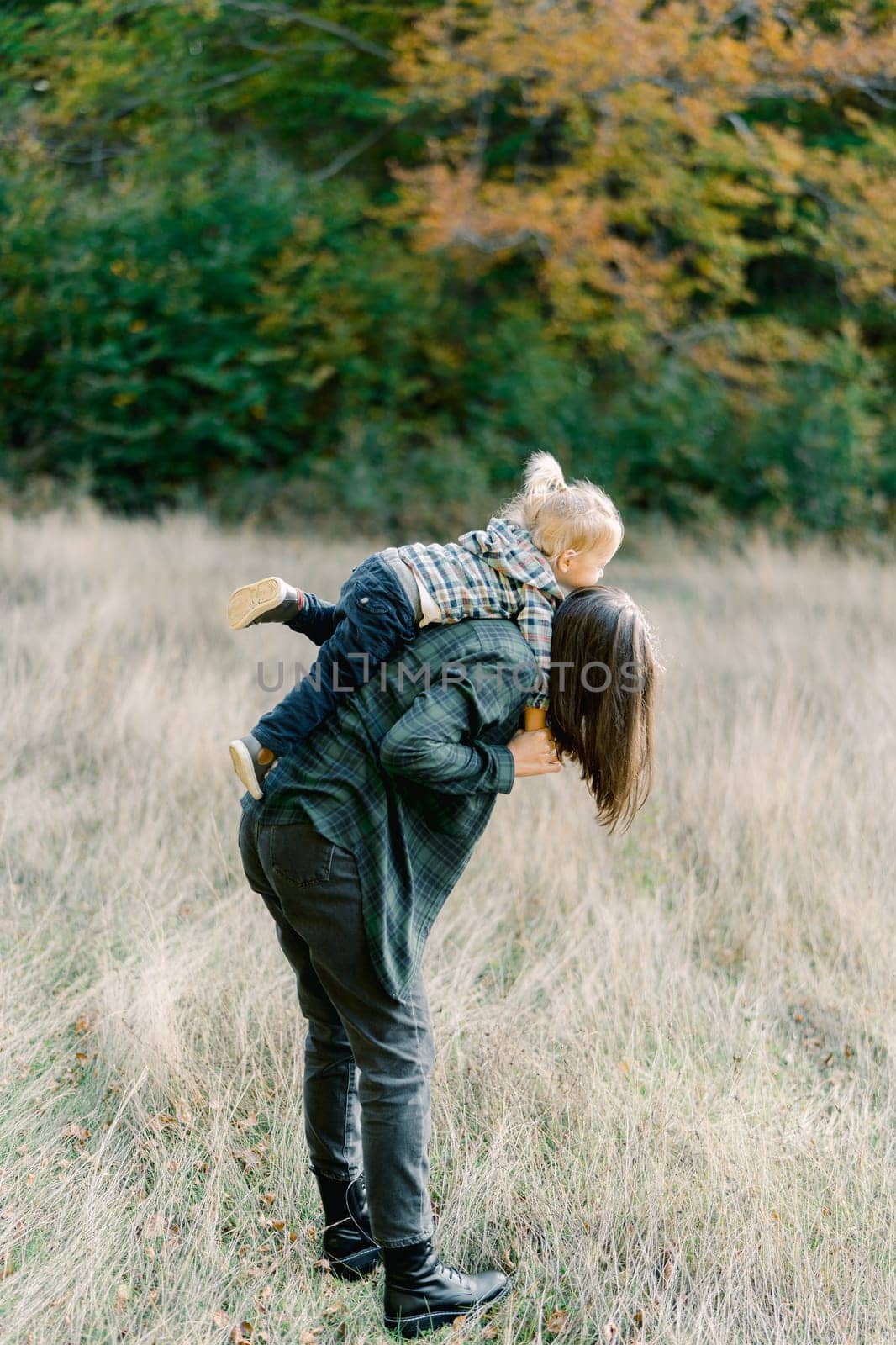 Mom with a little girl on her back leaned forward standing on the lawn. Side view. High quality photo