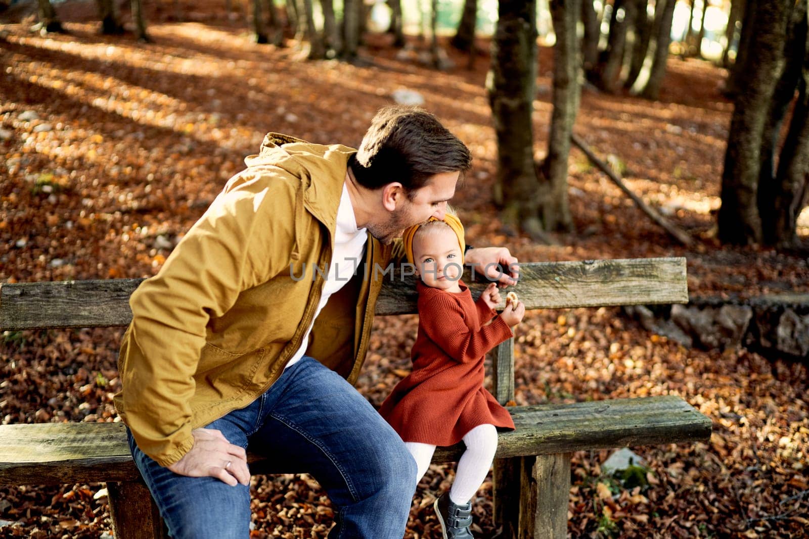 Dad wants to bite a bun in the hand of a little girl sitting with her on a bench in the autumn park. High quality photo
