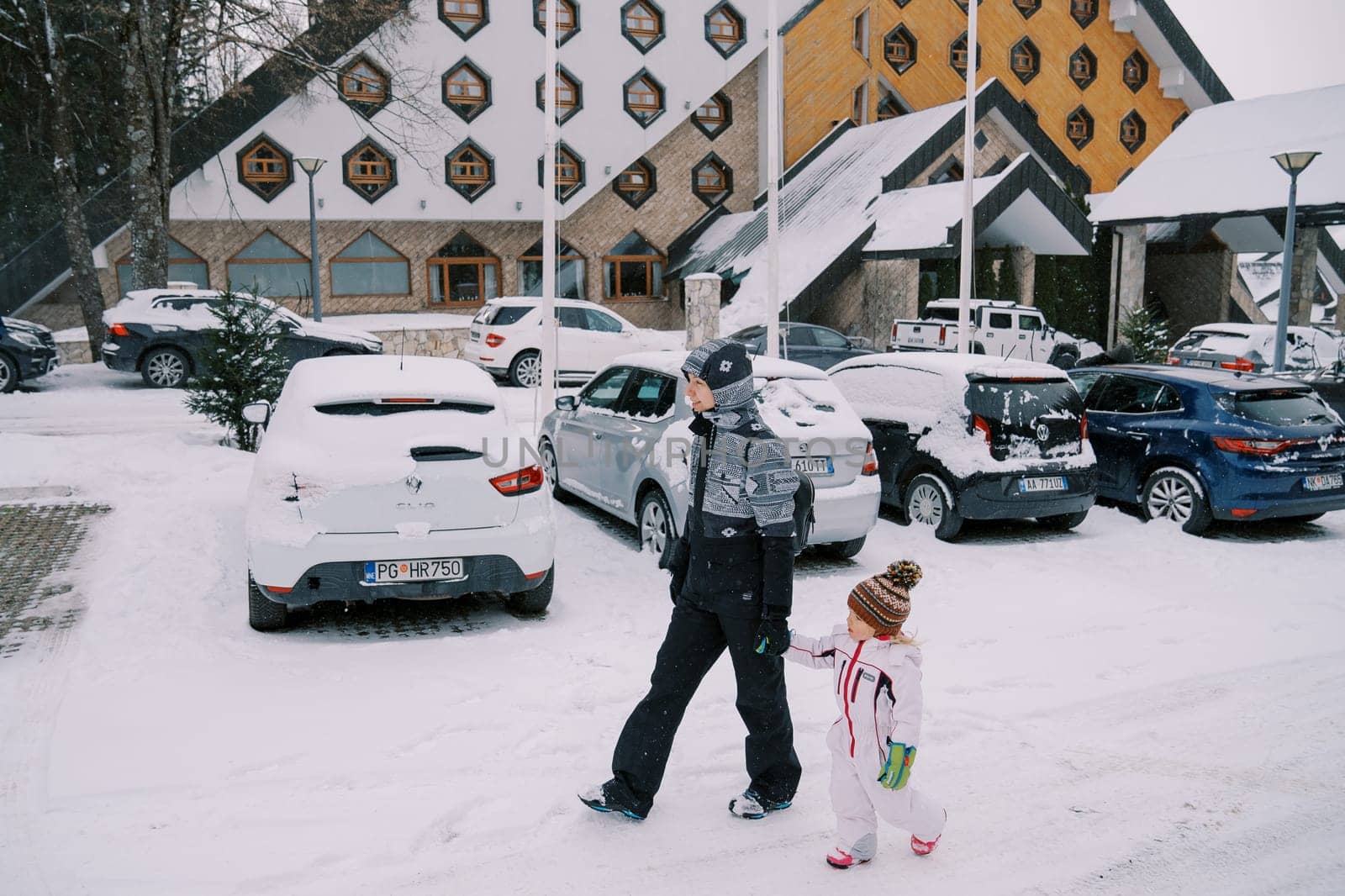 Mother and a little girl walk holding hands past cars parked outside a snow-covered hotel. Side view by Nadtochiy