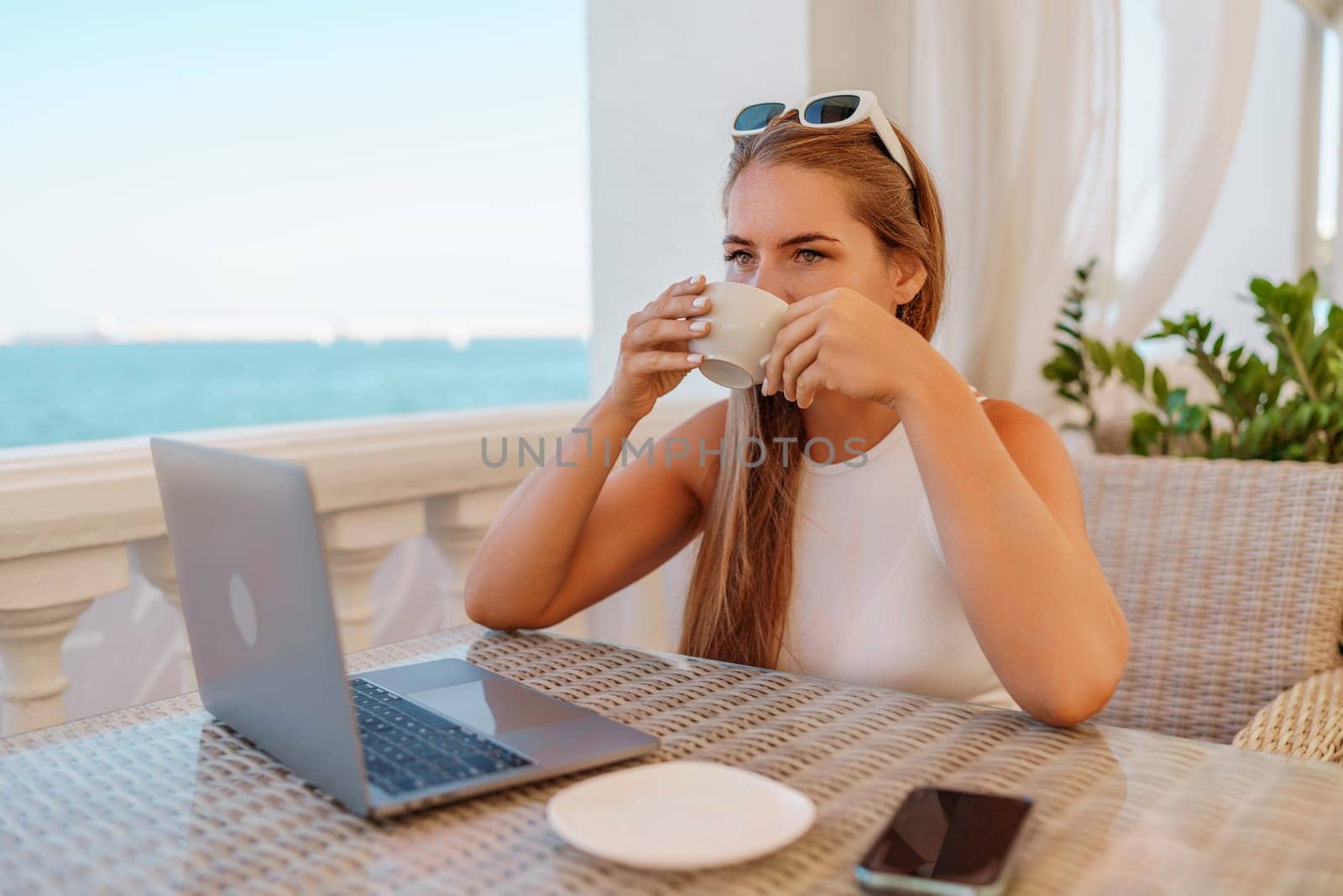 Woman coffee cafe laptop. Coffee break in cafe with sea view. Tranquil long haired woman drinking coffee in plant filled place. Woman sitting at a coffee shop with mobile phone drinking coffee. by Matiunina