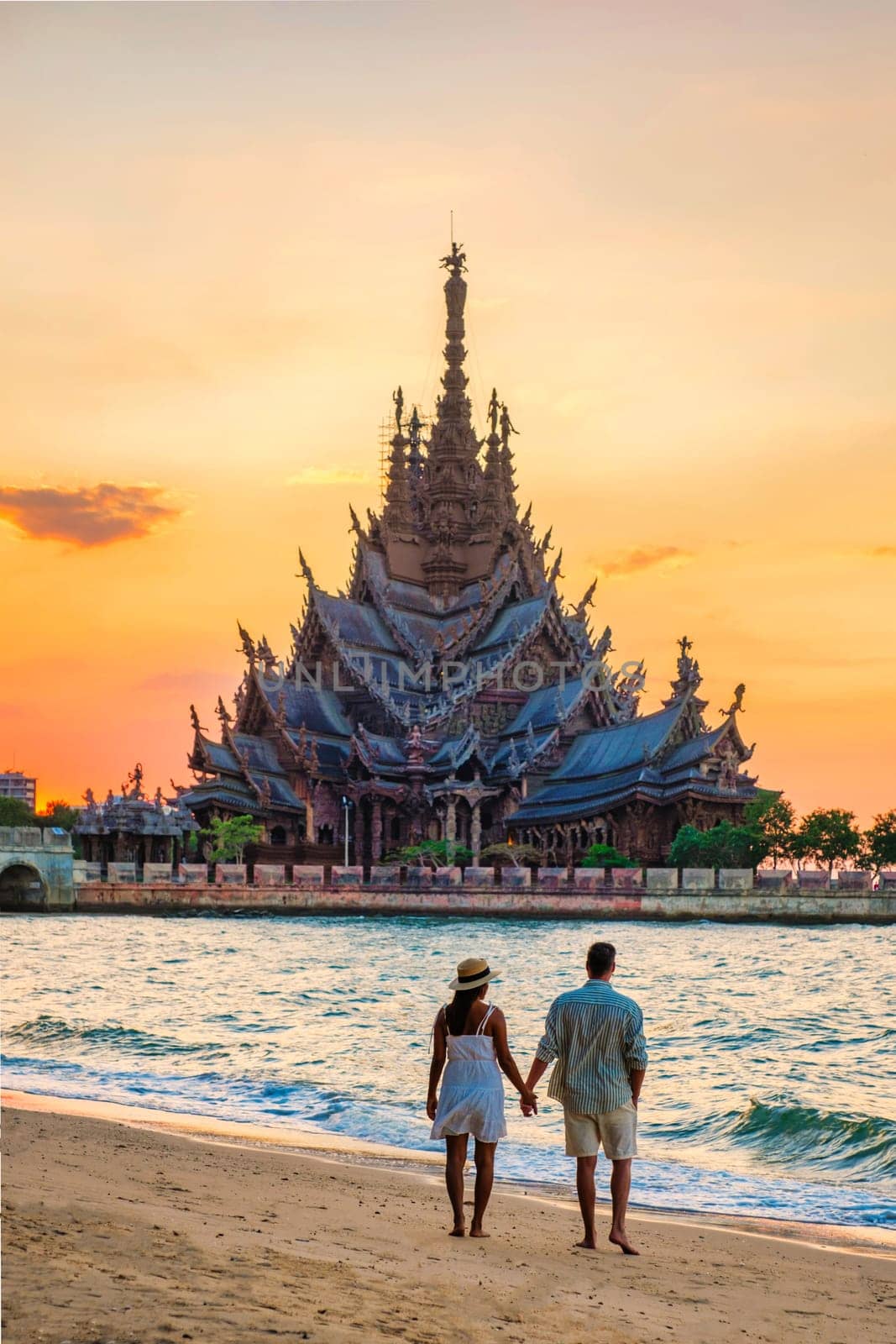 A diverse multiethnic couple of men and women visit The Sanctuary of Truth wooden temple in Pattaya Thailand at sunset on the beach looking at the wooden temple at dusk