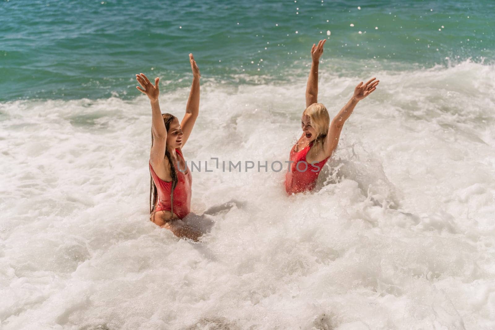 Women ocean play. Seaside, beach daytime, enjoying beach fun. Two women in red swimsuits enjoying themselves in the ocean waves and raising their hands up