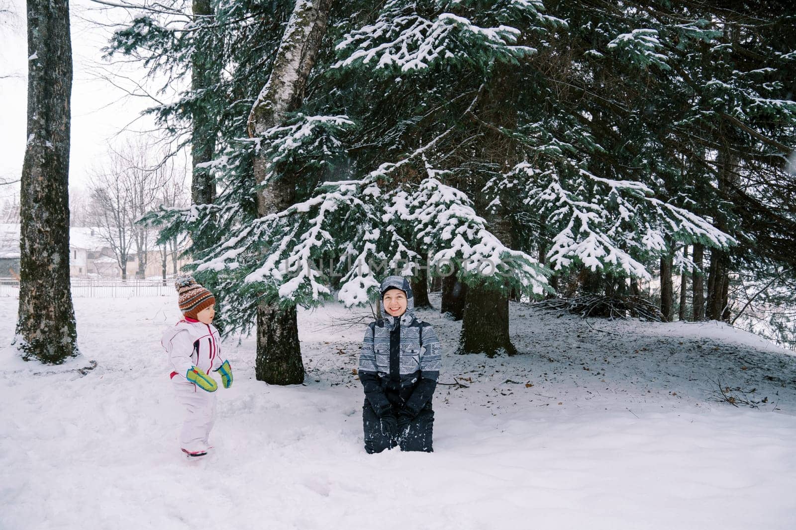 Little girl looks at her smiling mother sitting under a snow-covered spruce tree in a coniferous forest by Nadtochiy