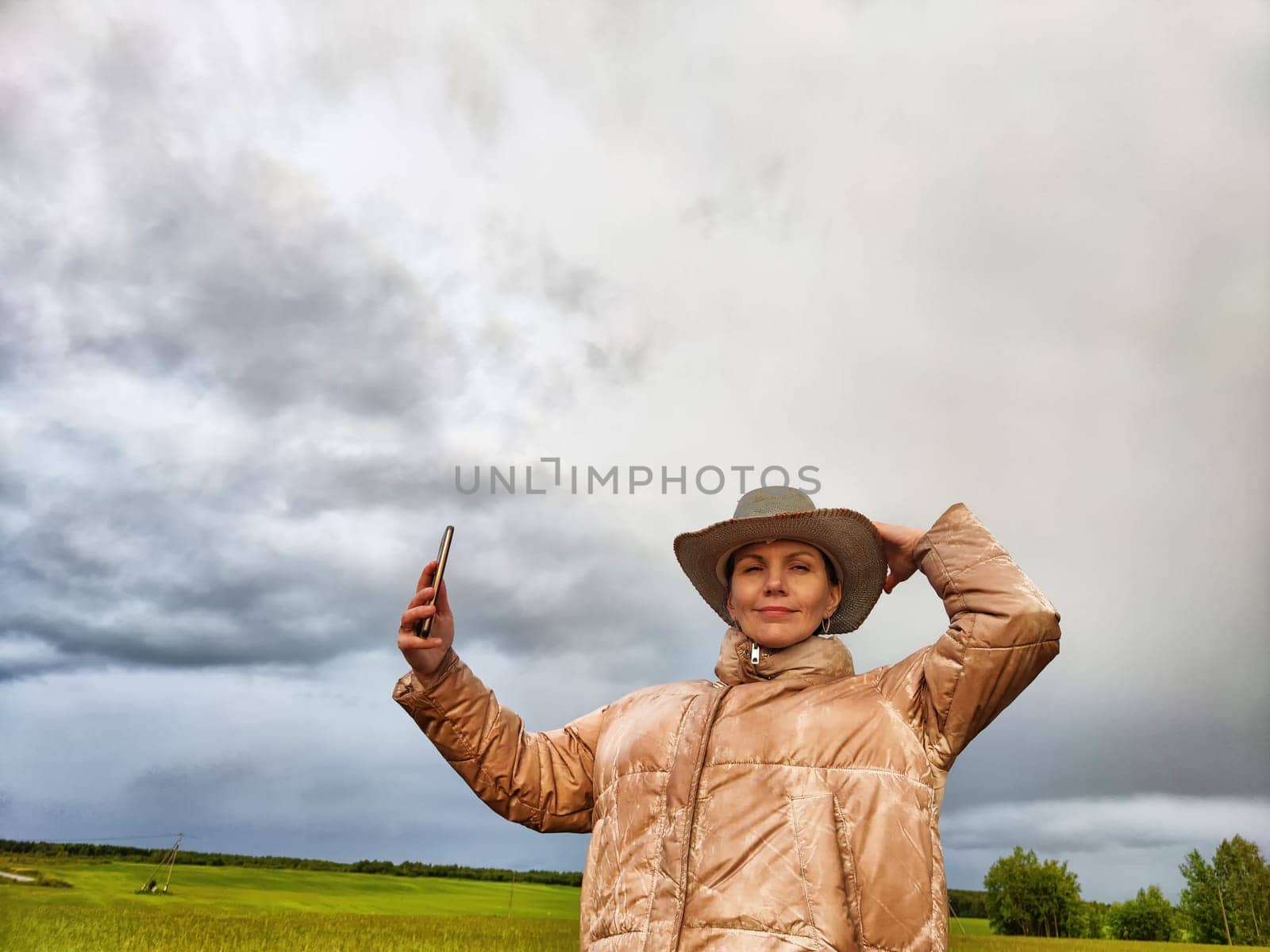 An adult girl in a field and with a stormy sky with clouds takes pictures of a rainbow and takes selfie in the rain. A woman having fun outdoors on rural and rustic nature