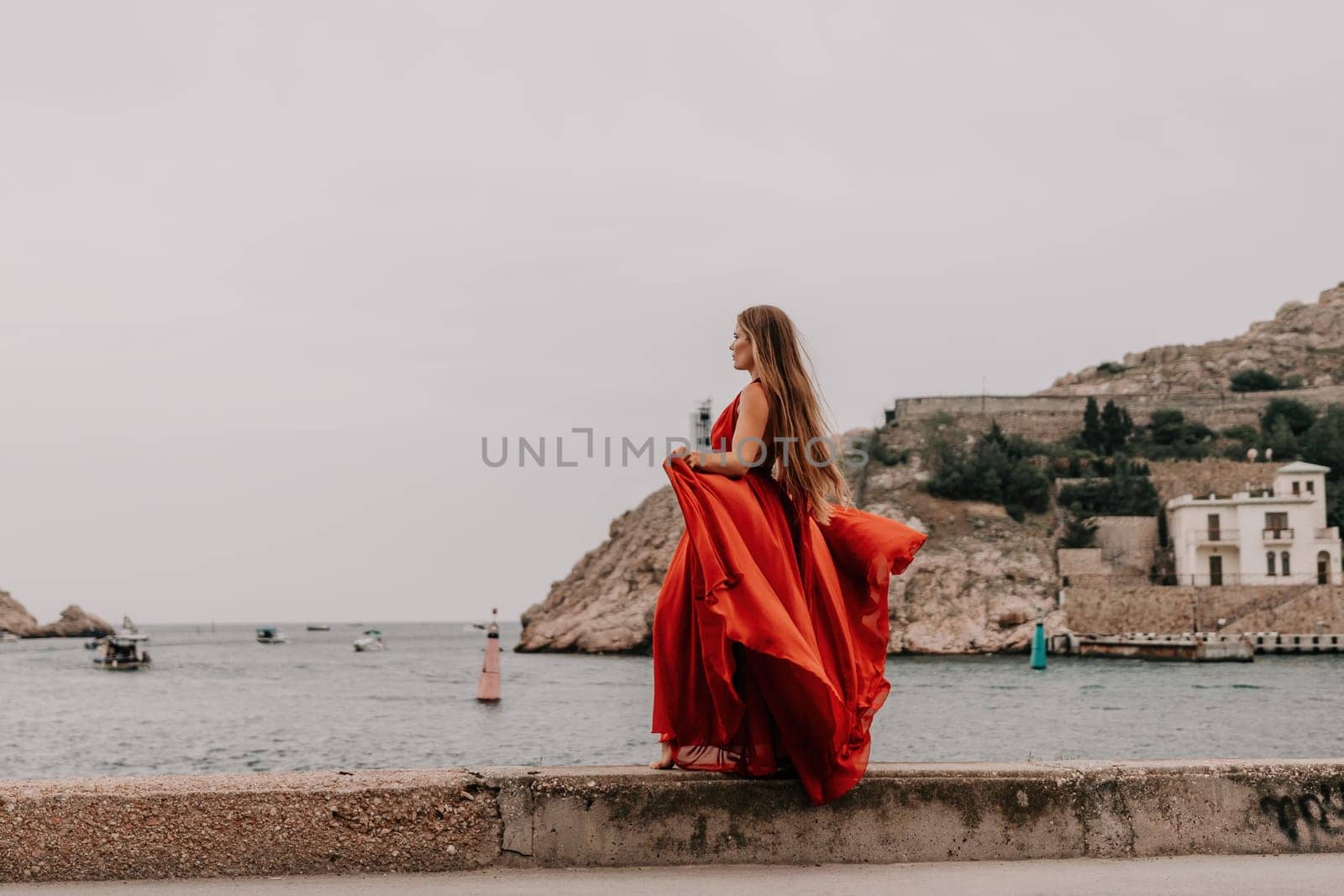 Side view a Young beautiful sensual woman in a red long dress posing on a volcanic rock high above the sea during sunset. Girl on the nature on overcast sky background. Fashion photo