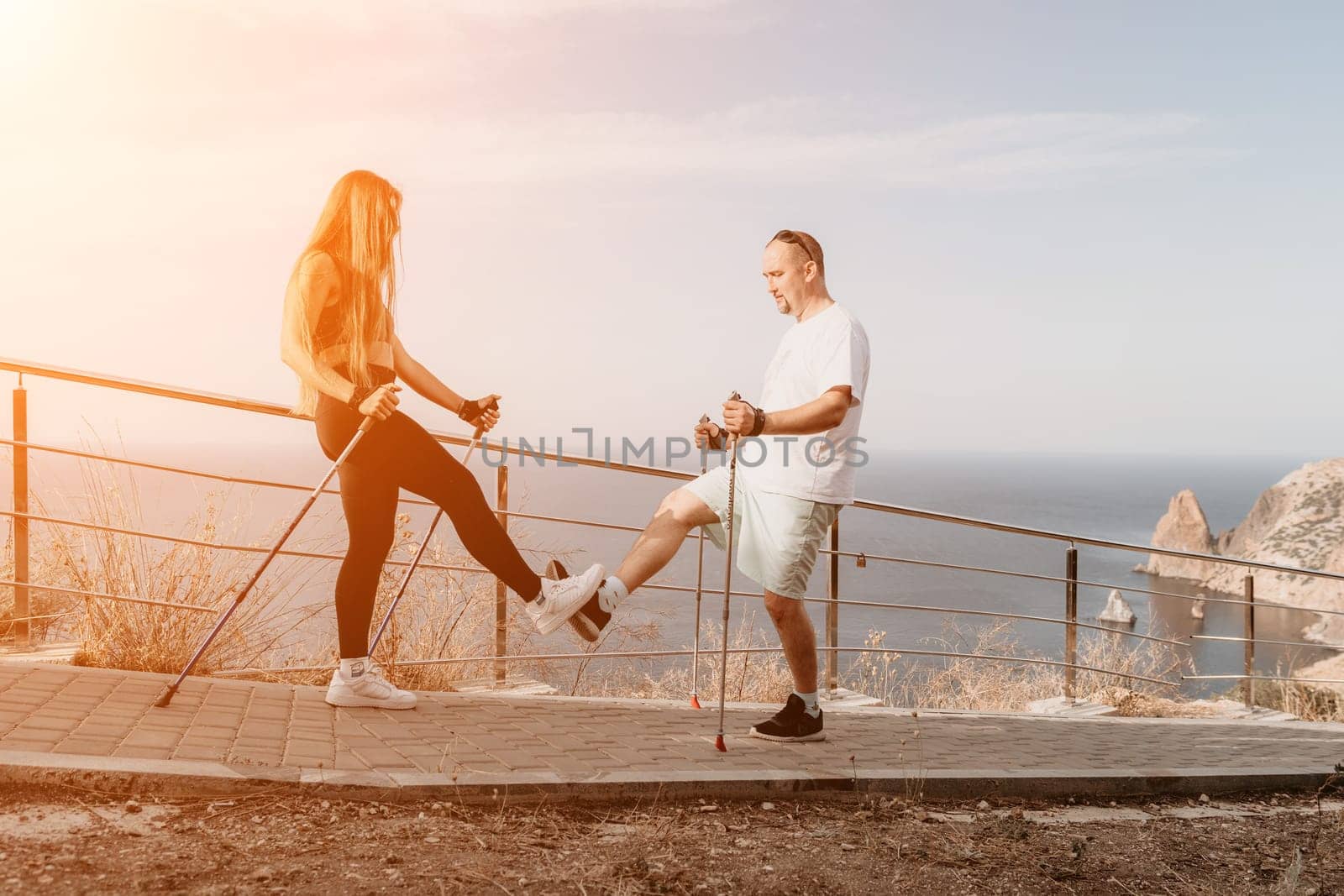 Happy Middle aged couple or friends practicing nordic walking in park near sea. Mature couple with trekking poles walking, practicing Nordic walking outdoors. Aging youthfully and sport concept by panophotograph