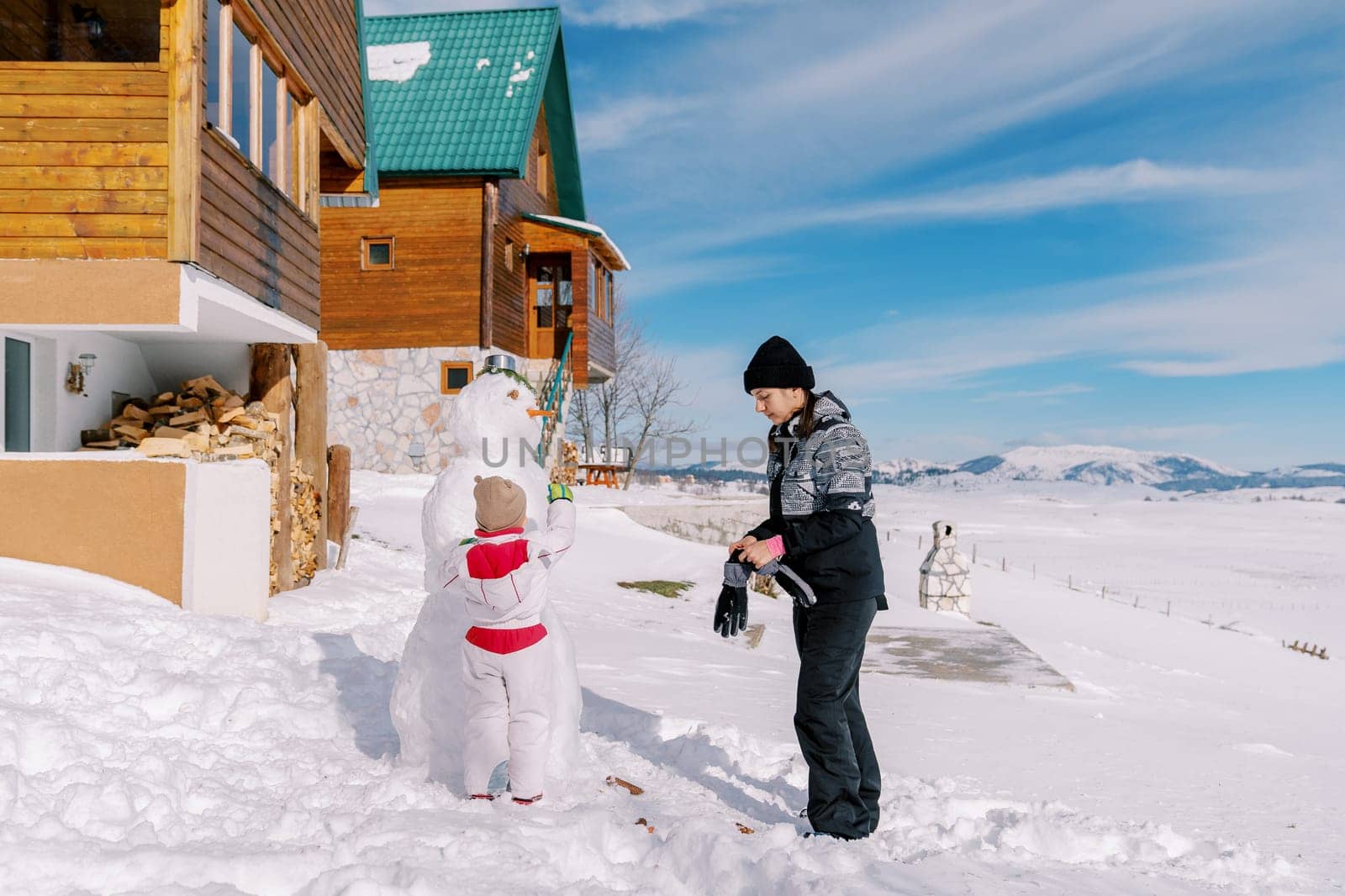 Little girl decorates a snowman near the cottage while standing next to her mother putting on mittens by Nadtochiy