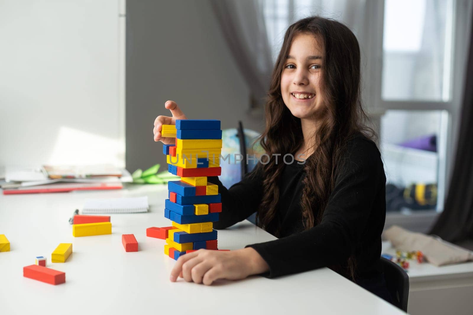 a little happy girl is playing the board game jenga at the table. Construction of a tower made of wooden cubes.