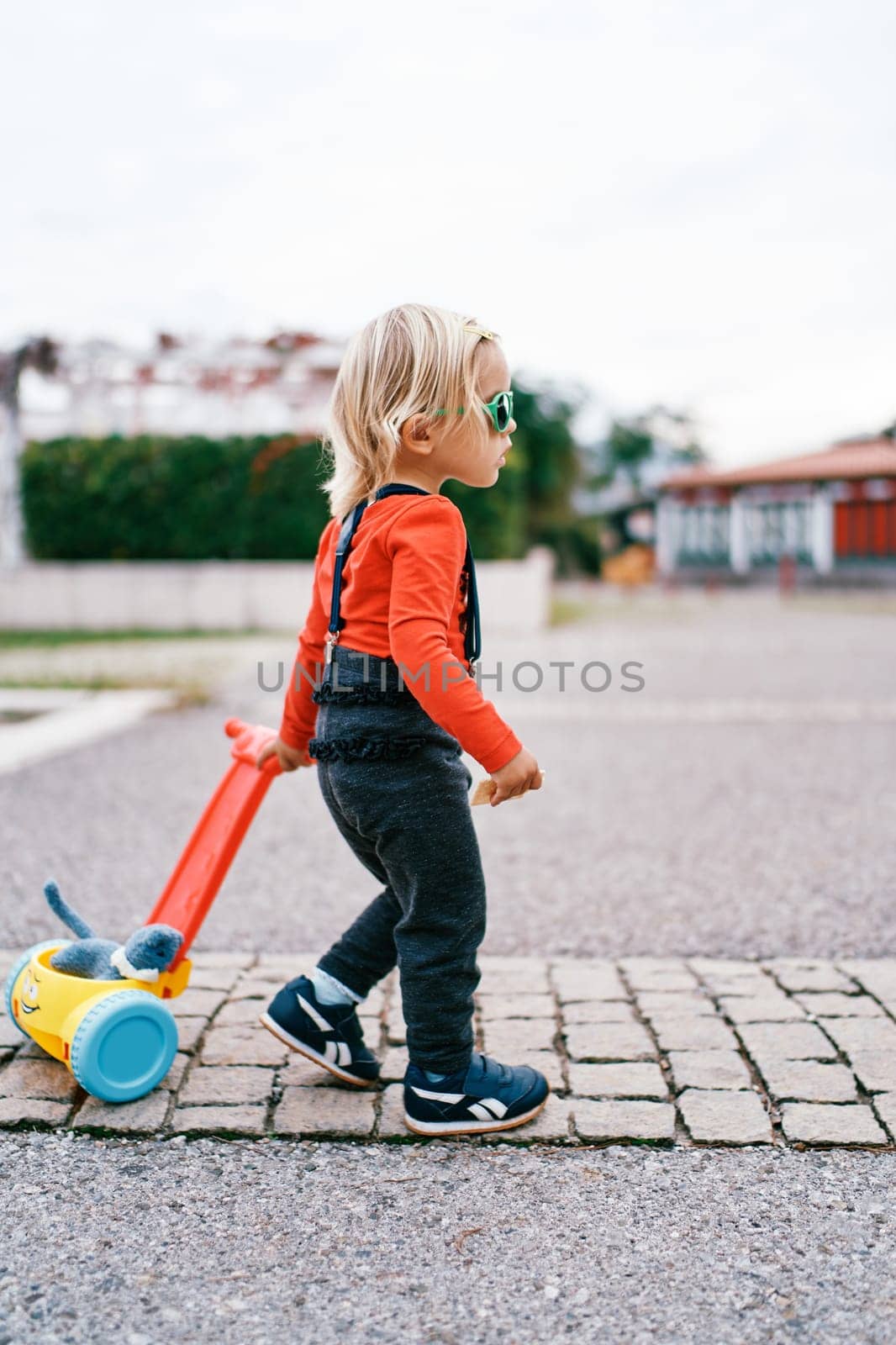 Little girl is carrying a toy cart with a soft toy on the paving stones. Side view. High quality photo