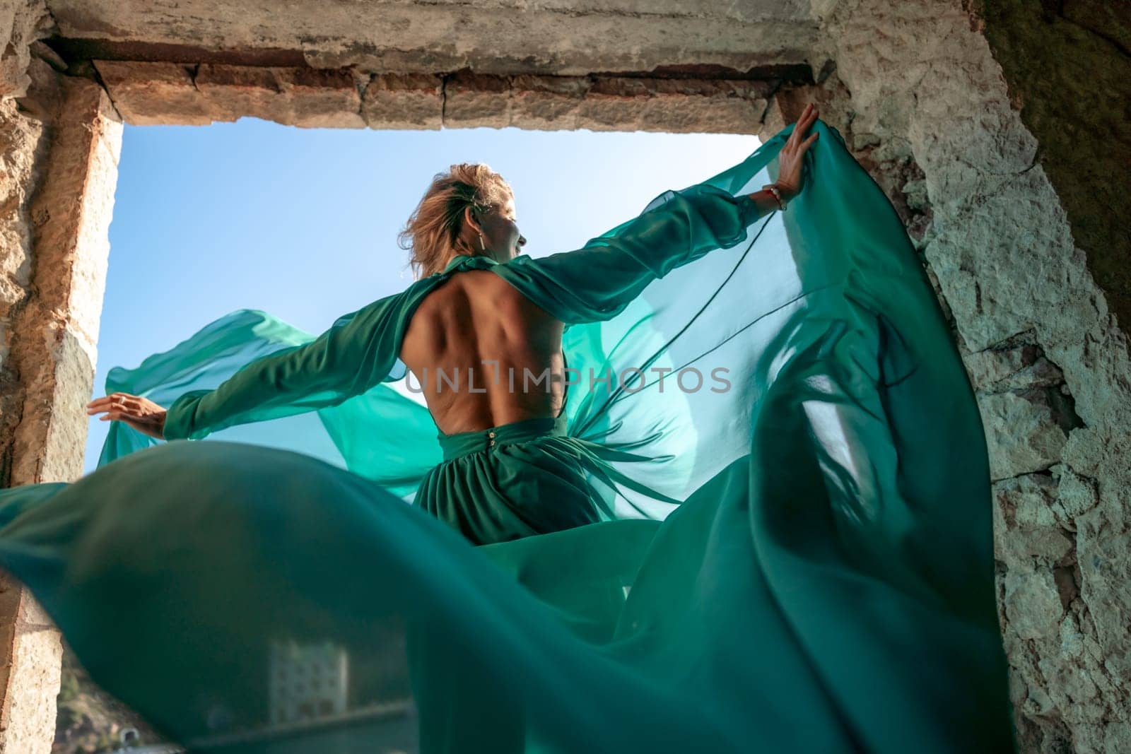 Rear view of a happy blonde woman in a long mint dress posing against the backdrop of the sea in an old building with columns. Girl in nature against the blue sky