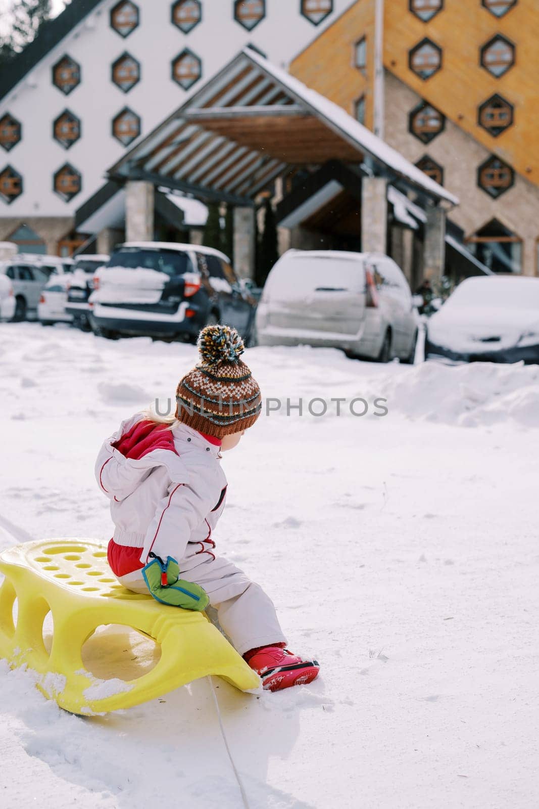 Small child sits on a sled in the snow near cars parked near the hotel. Back view. High quality photo