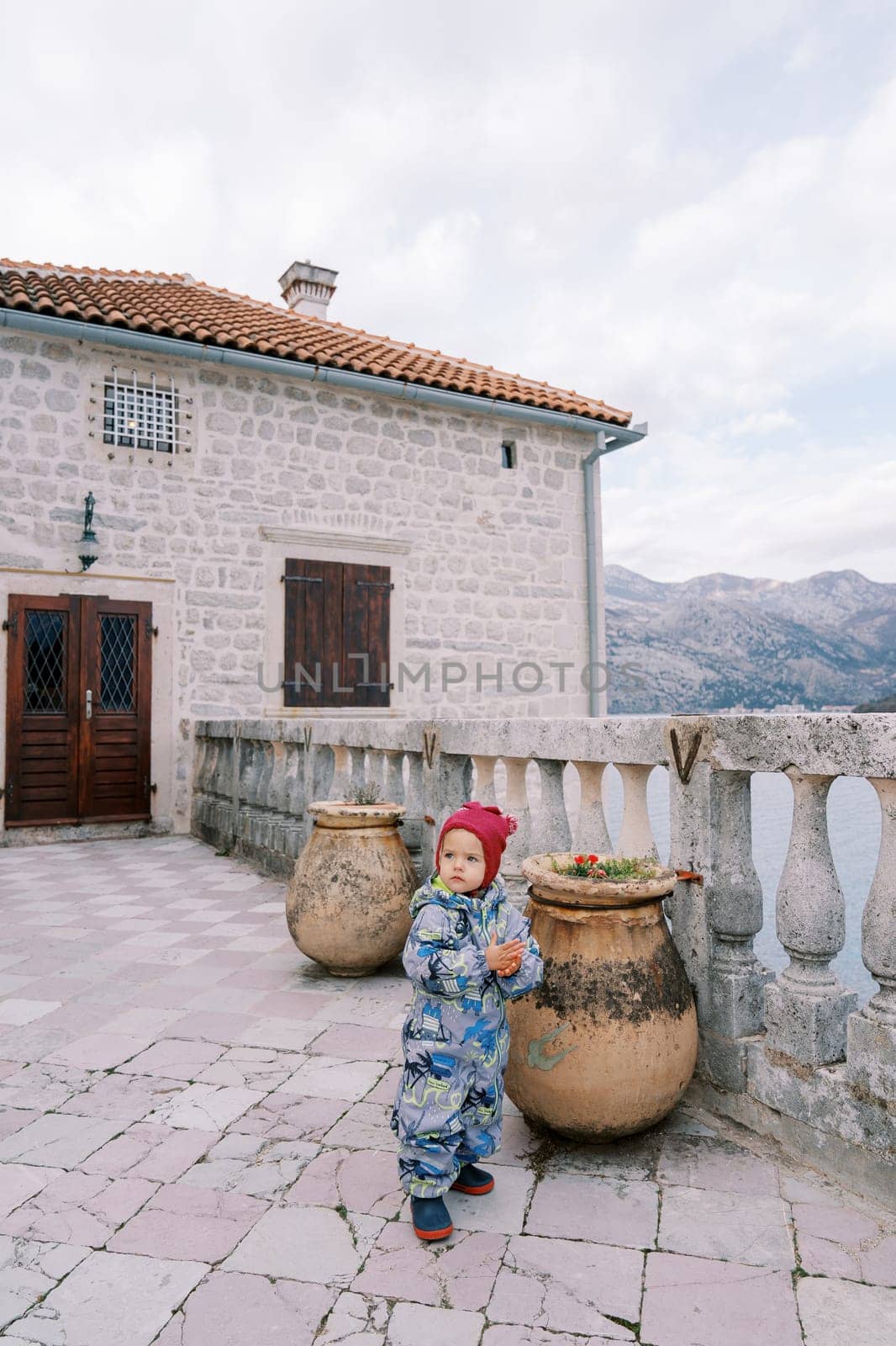 Little girl stands near a clay pot with flowers near a stone balustrade and looks away. High quality photo