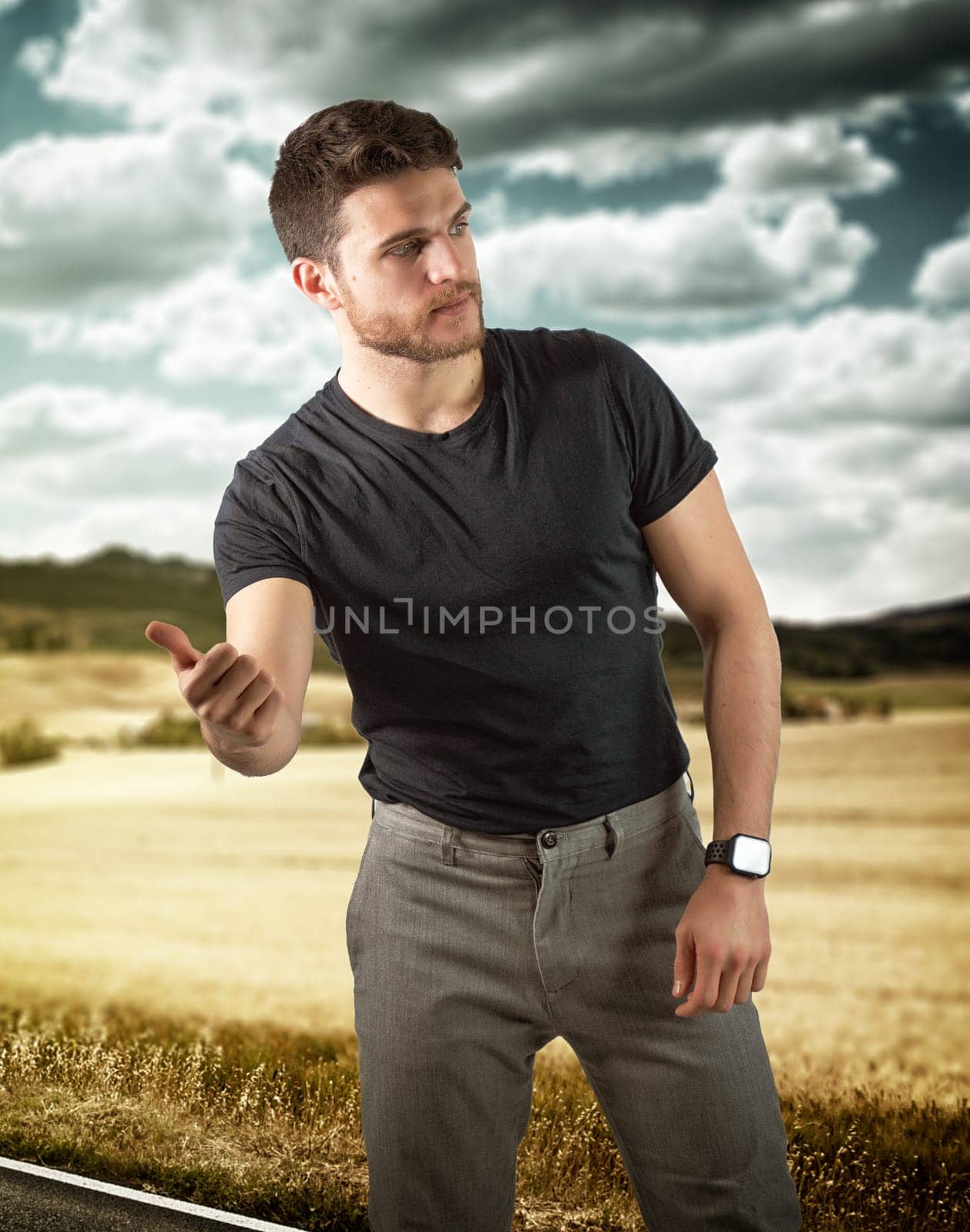 Handsome young man, a hitchhiker waiting for car on roadside in city, wearing black t-shirt
