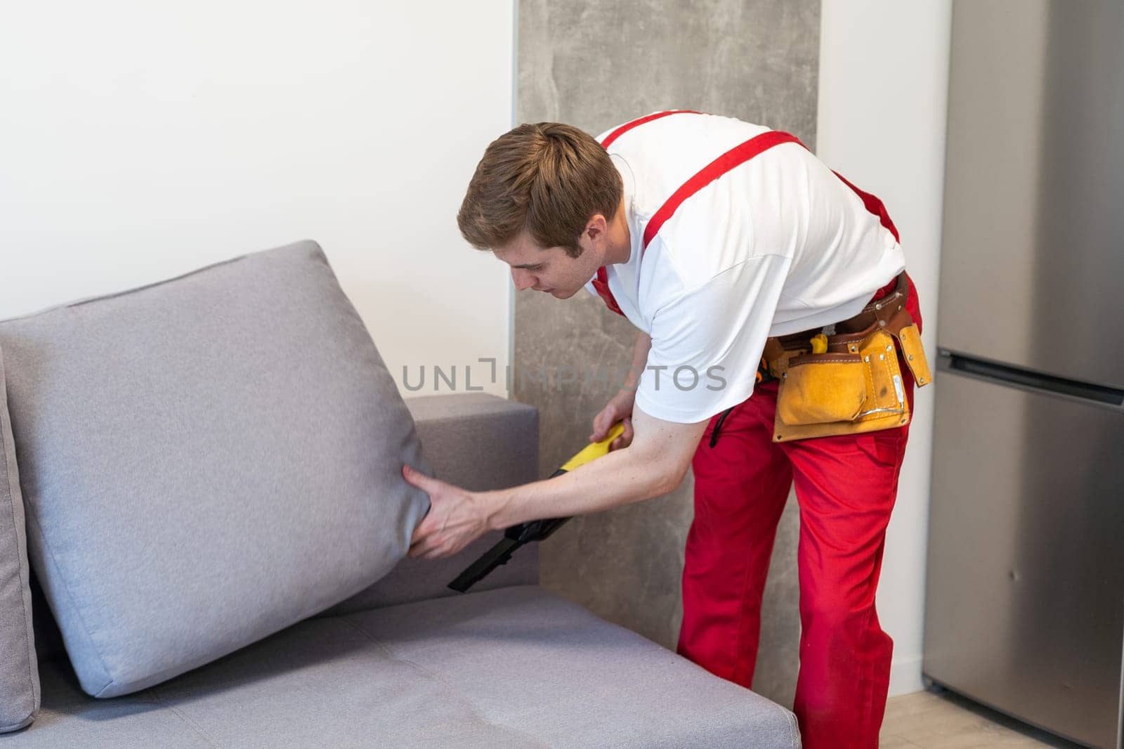 Rear View Of Young Male Worker Cleaning Sofa With Vacuum Cleaner.